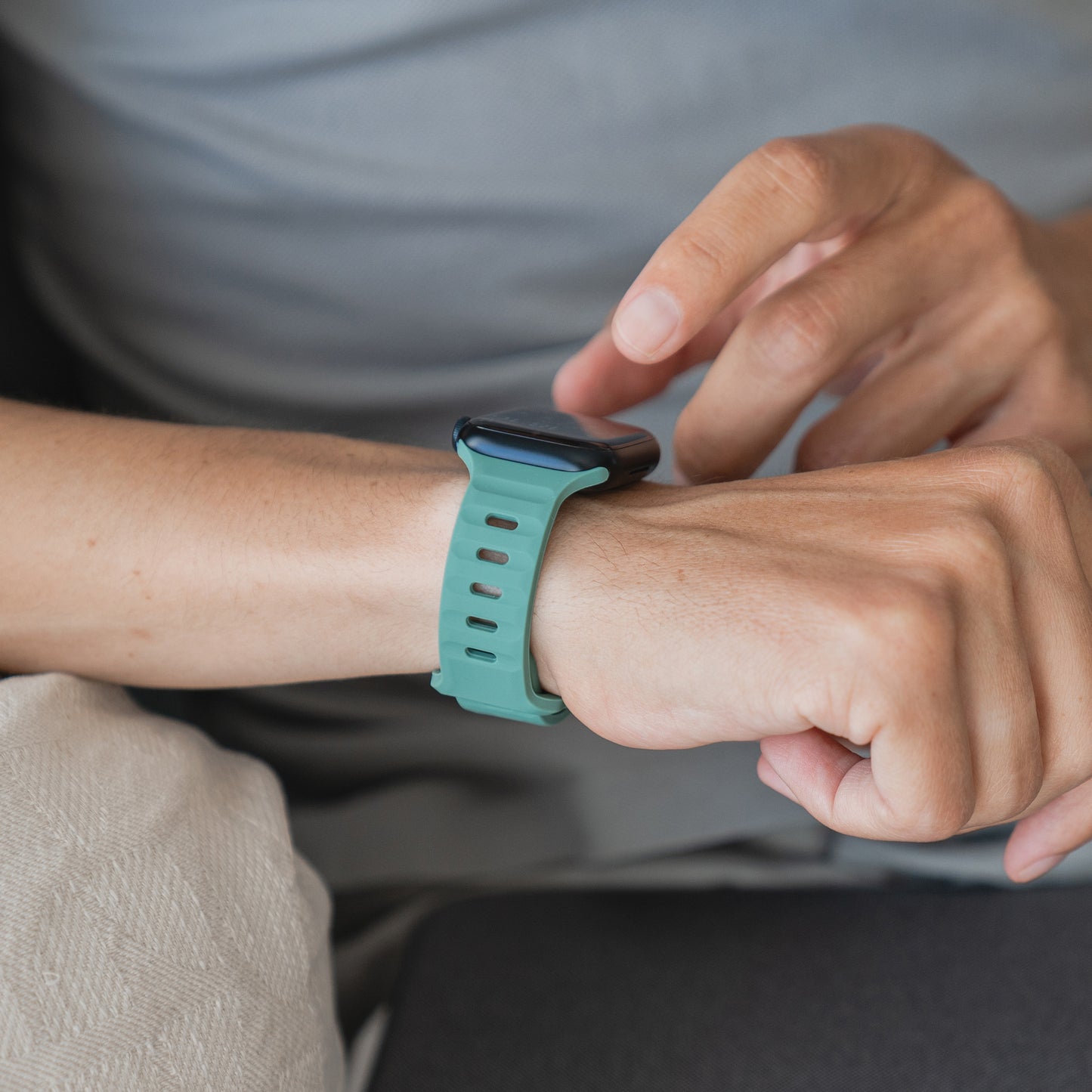 Eine Person in einem hellgrauen Hemd trägt ein Arktisband Apple Watch Silikonarmband „Active“ mit einem atmungsaktiven grünen Silikondesign am linken Handgelenk und interagiert mit der rechten Hand mit dem Gerät.