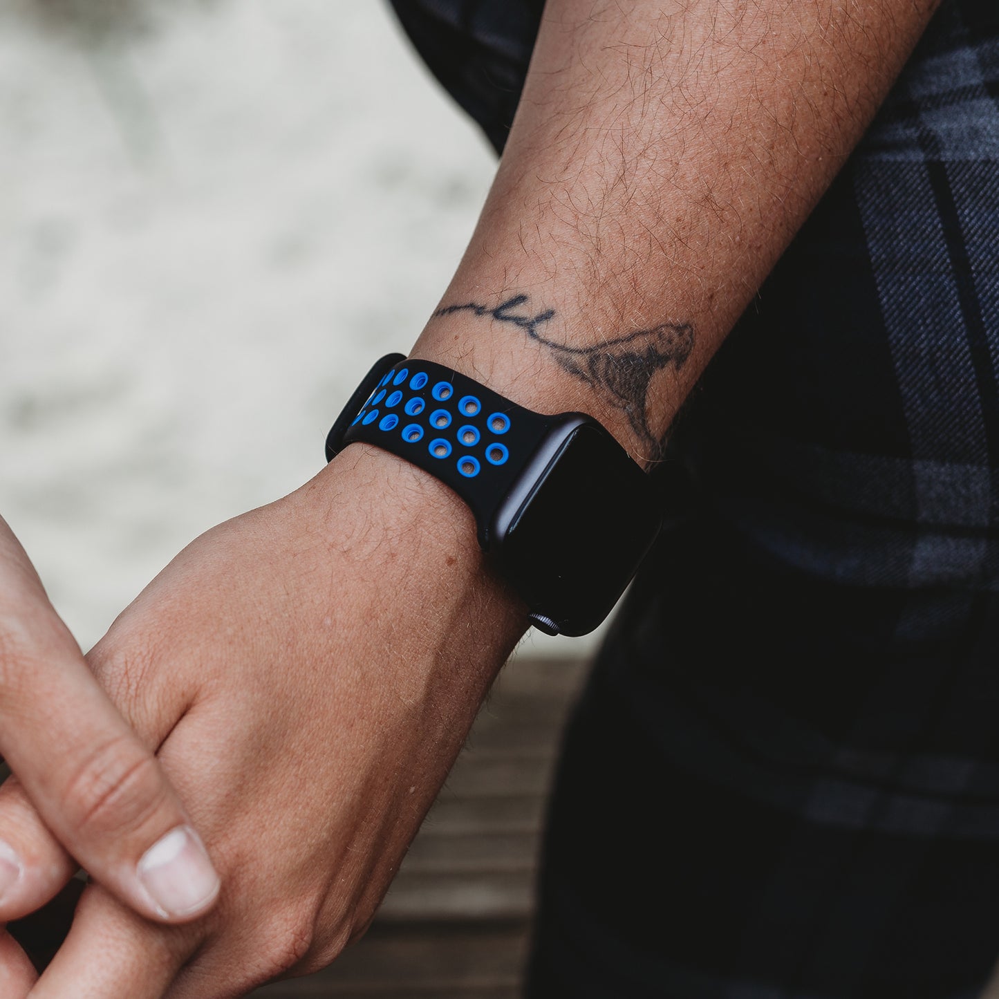 Eine Nahaufnahme zeigt ein Handgelenk mit dem arktisband Apple Watch Silikonarmband "Energy" von arktisband. Das schwarze Armband weist blaue Löcher auf. Die Person trägt ein Vogeltattoo auf dem Unterarm und trägt dunkel karierte Kleidung.