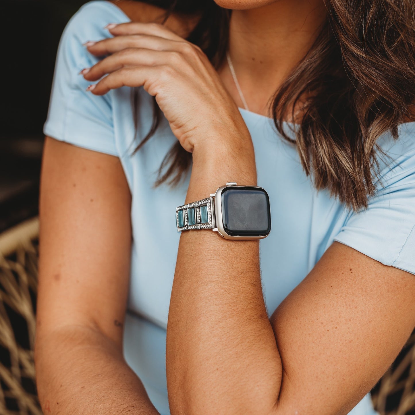 Eine Person mit mittellangem braunem Haar in einem hellblauen Kurzarmhemd verschränkt die Arme und zeigt ein arktisband Apple Watch Gliederarmband „Senorita“ von arktisband, mit silbernen und türkisfarbenen Perlen.