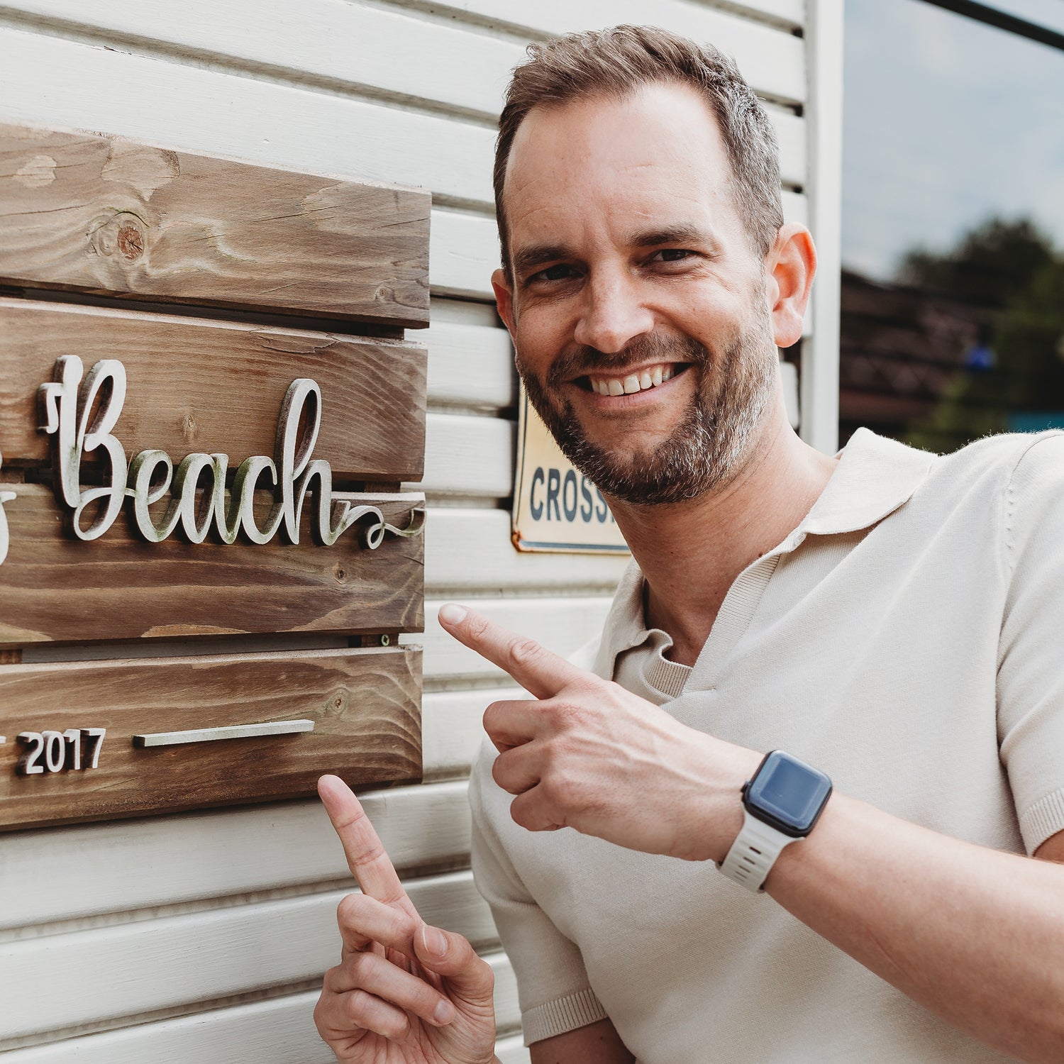 Ein Mann mit kurzen Haaren und Bart lächelt und zeigt auf ein Holzschild mit der Aufschrift „Beach“. Er trägt ein helles Poloshirt und das arktisband Apple Watch Silikonarmband „Active“ von arktisband. Im Hintergrund ist eine helle Holzverkleidung zu sehen.