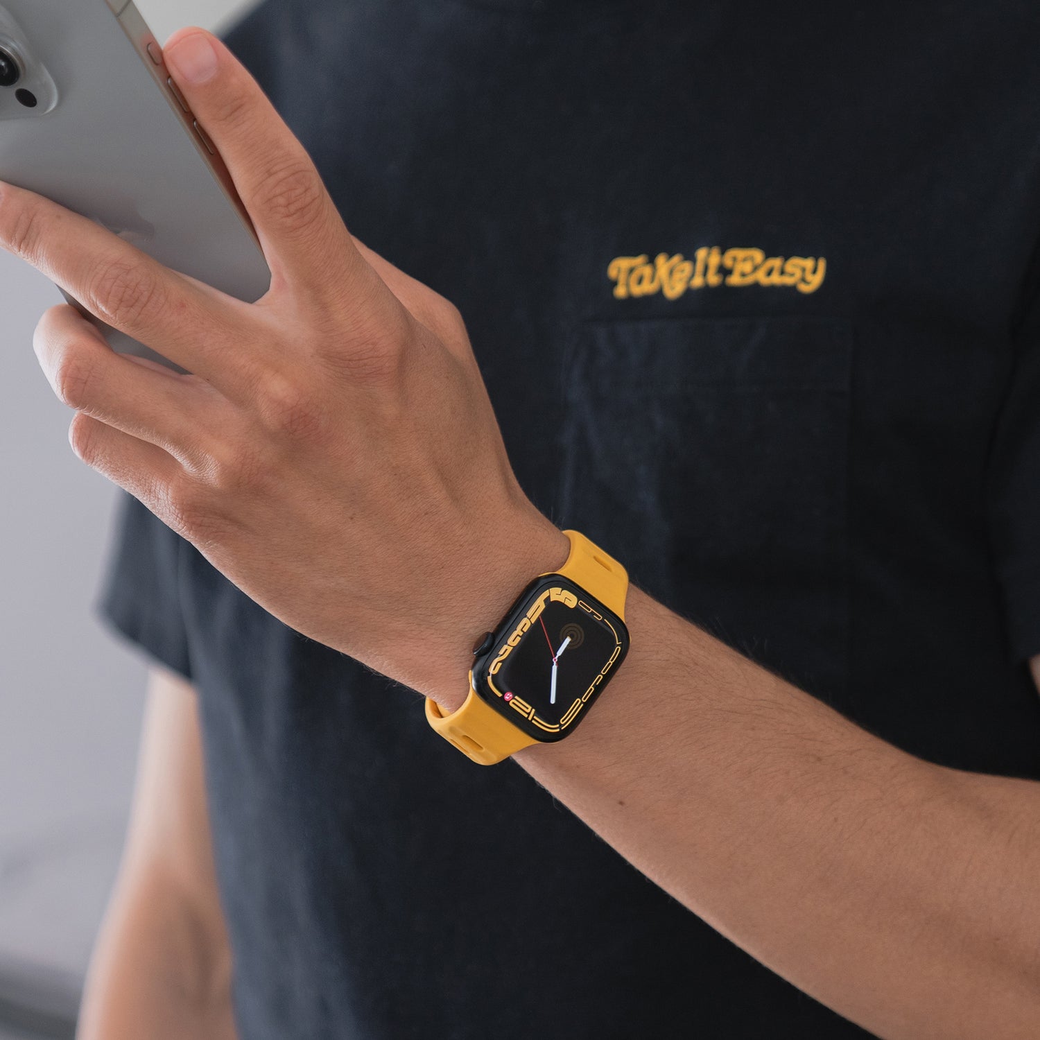 Eine Person in einem schwarzen T-Shirt mit „Take It Easy“ auf der Brust hält ein Smartphone und trägt neben einer Apple Watch Ultra ein Arktisband Apple Watch Silikonarmband „Active“, bekannt für seine Atmungsaktivität, am Handgelenk.