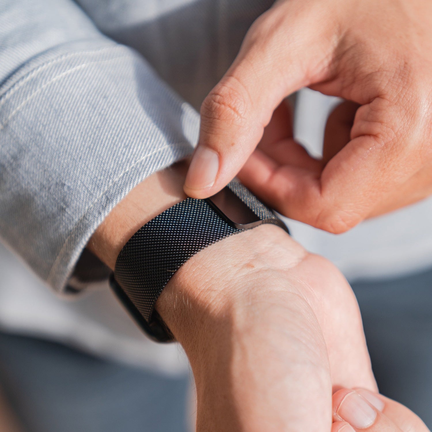 Eine Person befestigt ein schwarzes Arktisband Apple Watch Milanaise Loop Armband am linken Handgelenk und trägt ein helles Langarmshirt. Der Fokus liegt auf den Zeigern und der Apple Watch mit ihrem eleganten Mesh-Armband.