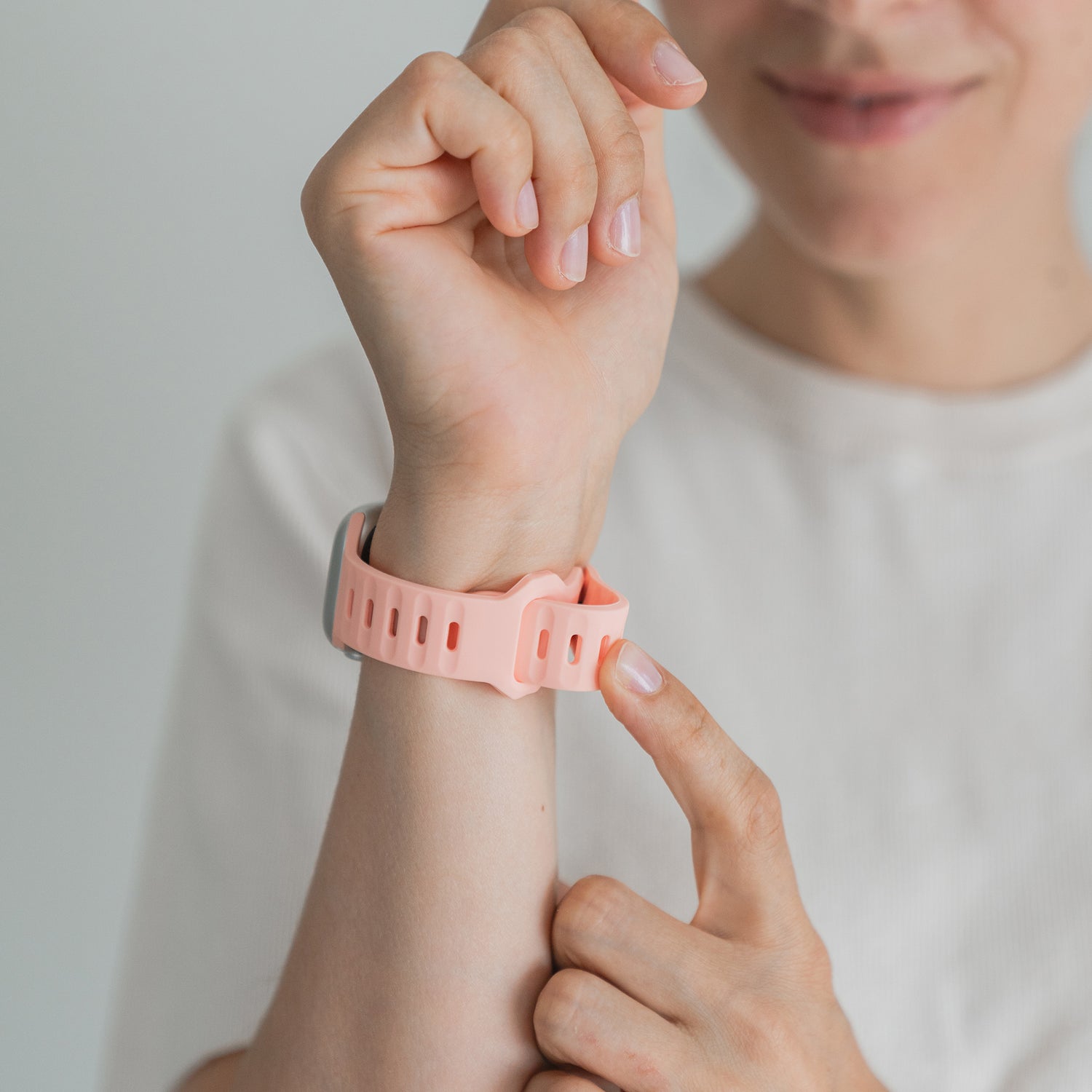 Eine Person im weißen Hemd zeigt auf ein rosa Arktisband Apple Watch Silikonarmband „Active“ an ihrem Handgelenk und lächelt leicht, wobei nur die untere Hälfte ihres Gesichts zu sehen ist.