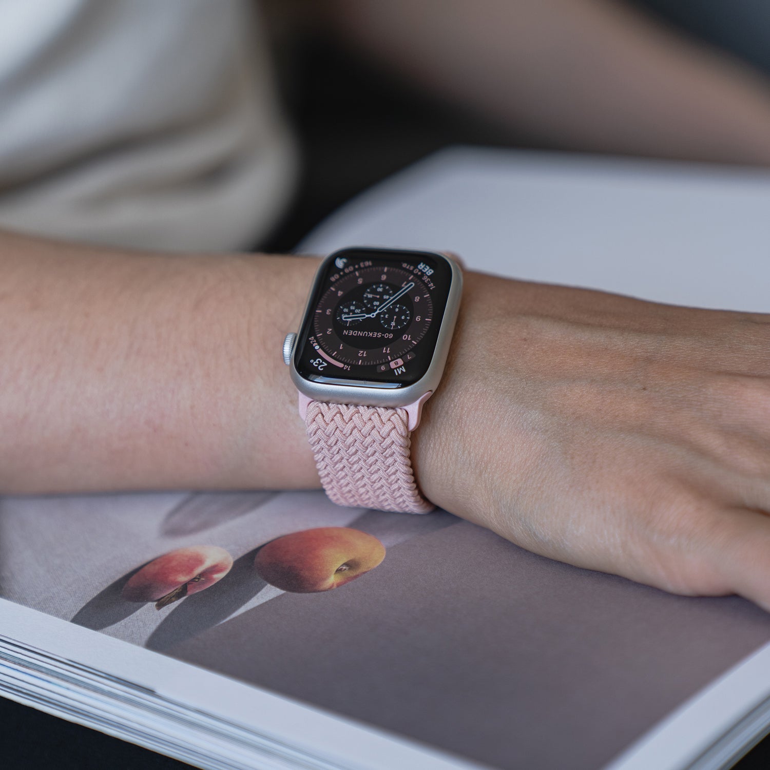 Eine Person, die ein arktisband Geflochtenes Apple Watch Armband „Casual“ mit Magnetverschluss in Pink trägt, legt ihre Hand auf ein aufgeschlagenes Magazin mit Pfirsichillustrationen.