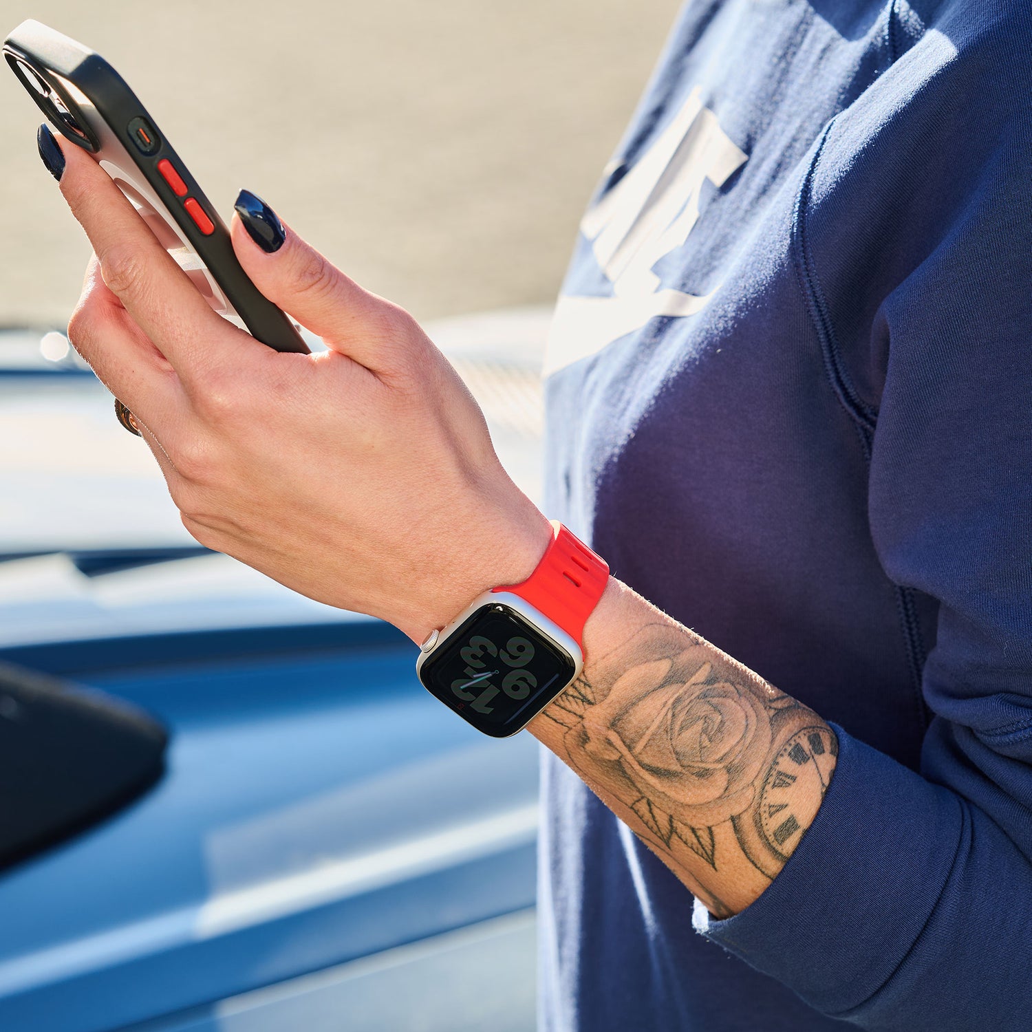 Eine Person in einem blauen Sweatshirt und einem Arktisband Apple Watch Silikonarmband „Active“ hält ein Smartphone in der Hand. Auf ihrem Unterarm sind ein Rosentattoo, dunkler Nagellack und im Hintergrund ein blaues Auto zu sehen.