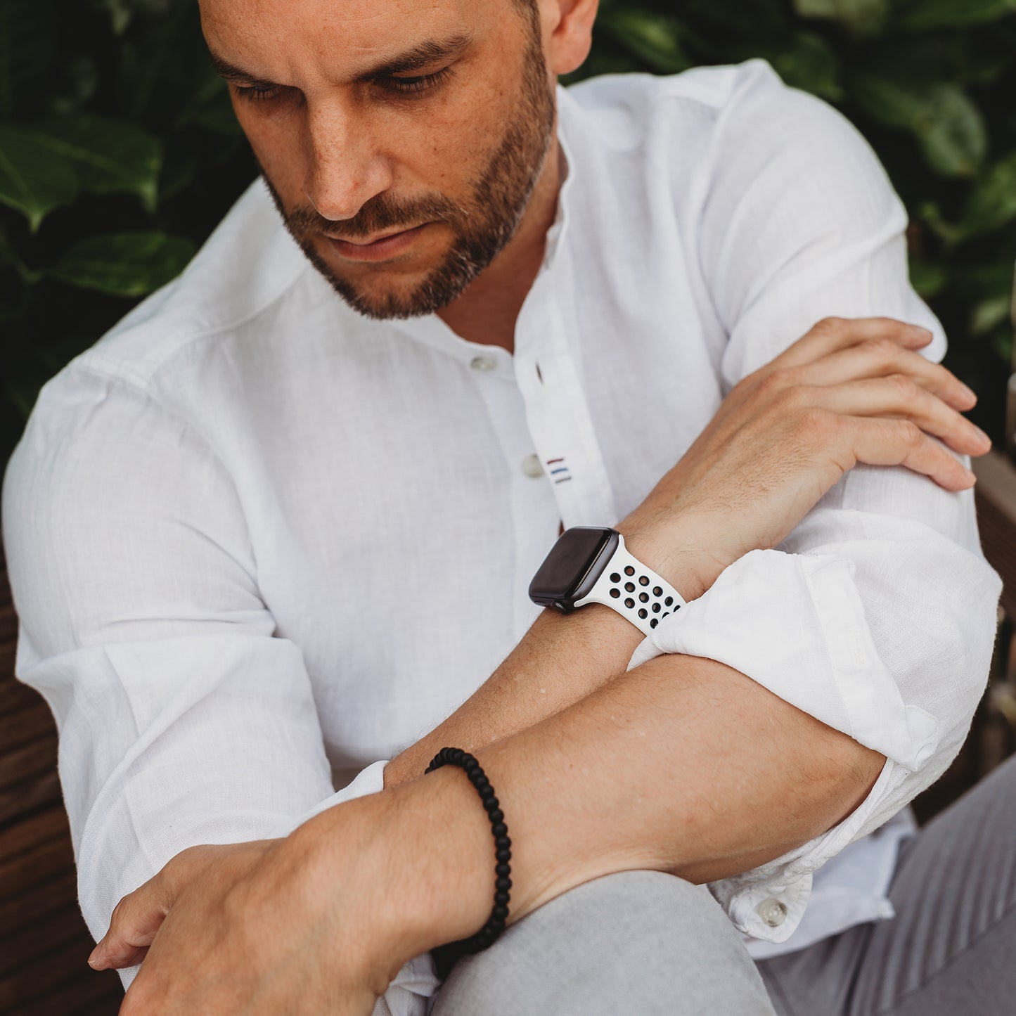 Ein Mann in einem weißen Button-Down-Hemd, der ein arktisband Apple Watch Silikonarmband „Energy“ von arktisband und ein schwarzes Perlenarmband trägt, sitzt mit verschränkten Armen da und blickt nachdenklich auf das üppige grüne Laub hinunter.