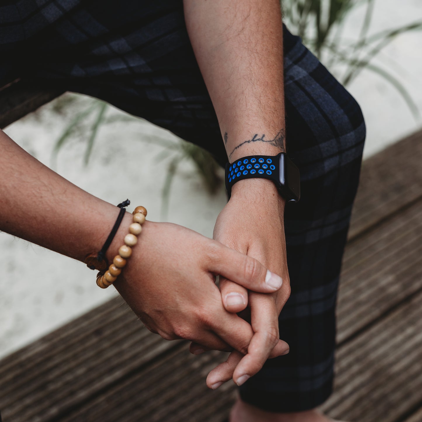 Eine Person mit einem Arktisband Apple Watch Silikonarmband „Energy“ und einem Perlenarmband sitzt in dunkelkarierten Hosen mit gefalteten Händen auf Holzstufen, im Hintergrund sind grüne Pflanzen zu sehen.