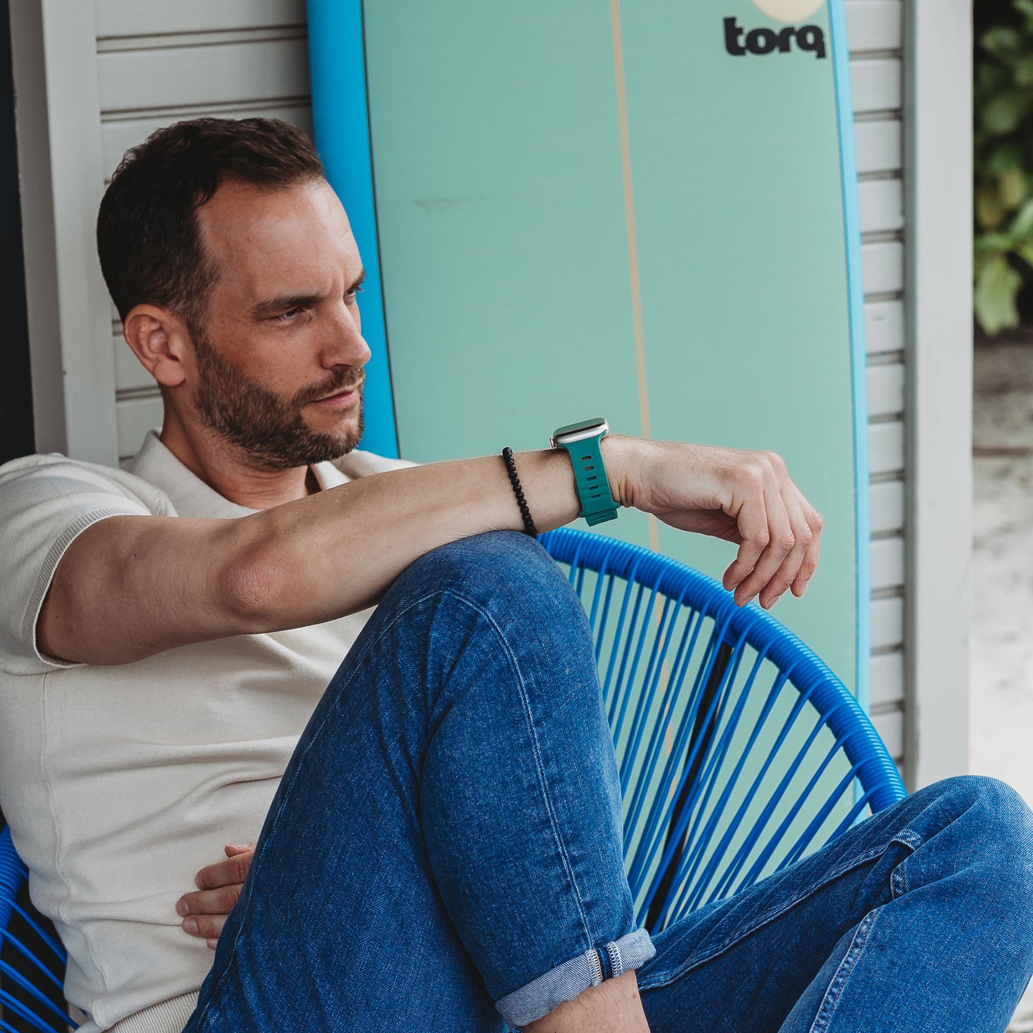 Ein Mann in weißem Hemd und Blue Jeans sitzt auf einem blauen Stuhl und überprüft sein Handgelenk mit einem Arktisband Apple Watch Silikonarmband „Active“. Im Hintergrund steht ein türkisfarbenes Surfbrett.