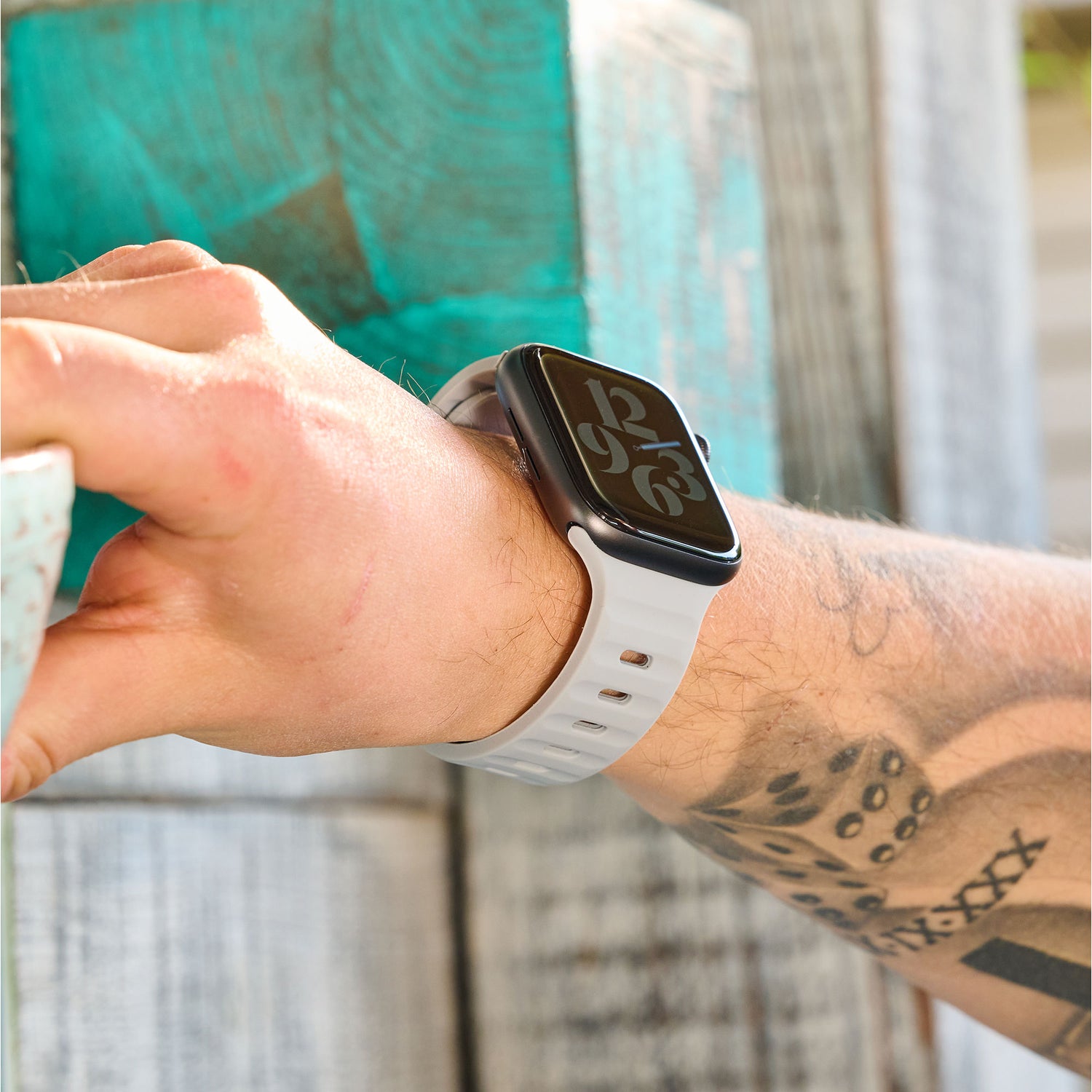 Eine Person mit Tattoos auf dem Unterarm trägt ein Arktisband Apple Watch Silikonarmband „Active“ in Weiß, das die Uhrzeit 12:16 anzeigt, und greift nach einer türkisfarbenen Holzoberfläche.