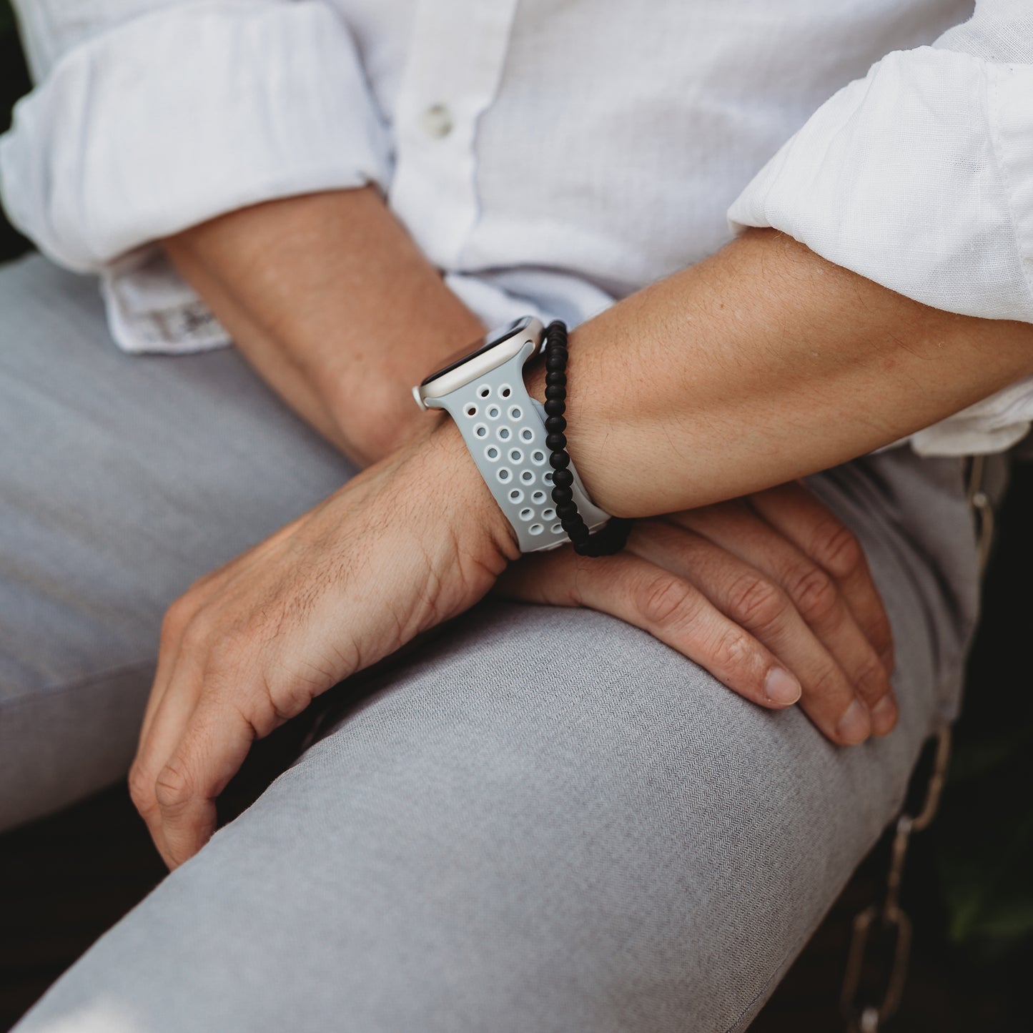 Eine Person in hellgrauer Hose und weißem Hemd sitzt mit den Händen auf den Knien und präsentiert das arktisband Apple Watch Silikonarmband „Energy“ von arktisband und ein schwarzes geflochtenes Armband an ihrem Handgelenk.