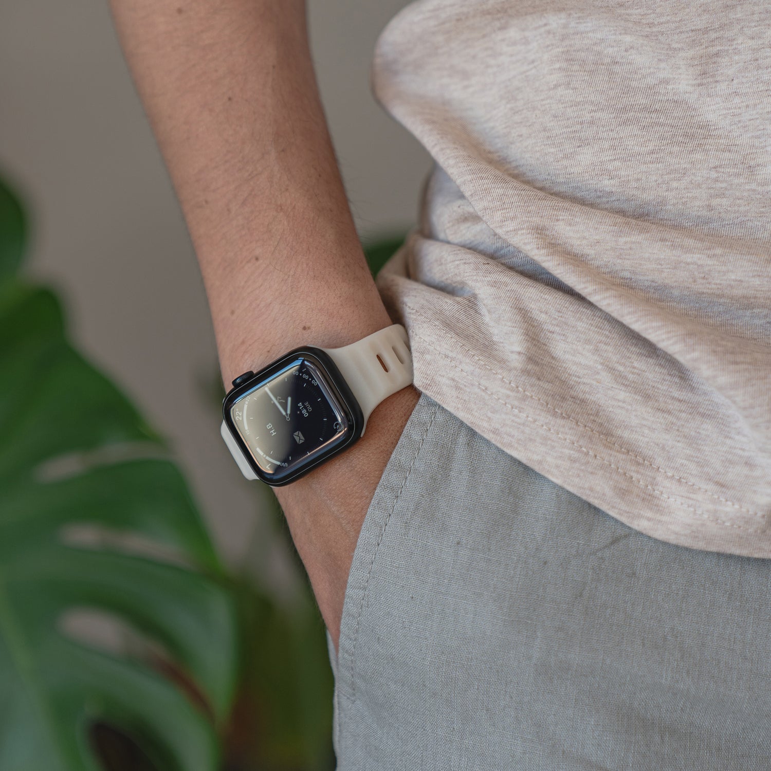 Eine Person in einem beigen T-Shirt und einer grauen Hose präsentiert ihre Apple Watch mit dem Arktisband „Active“-Silikonarmband, während ein großes grünes Blatt im Hintergrund für eine sportliche Atmosphäre sorgt.