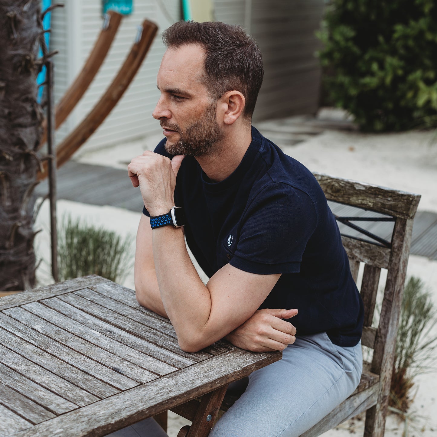 Ein Mann mit kurzen Haaren und Bart sitzt an einem Holztisch im Freien und trägt das arktisband Apple Watch Silikonarmband "Energy" von arktisband. Er legt seinen Arm auf den Tisch und blickt zur Seite, im Hintergrund sind Sand und Grün zu sehen.