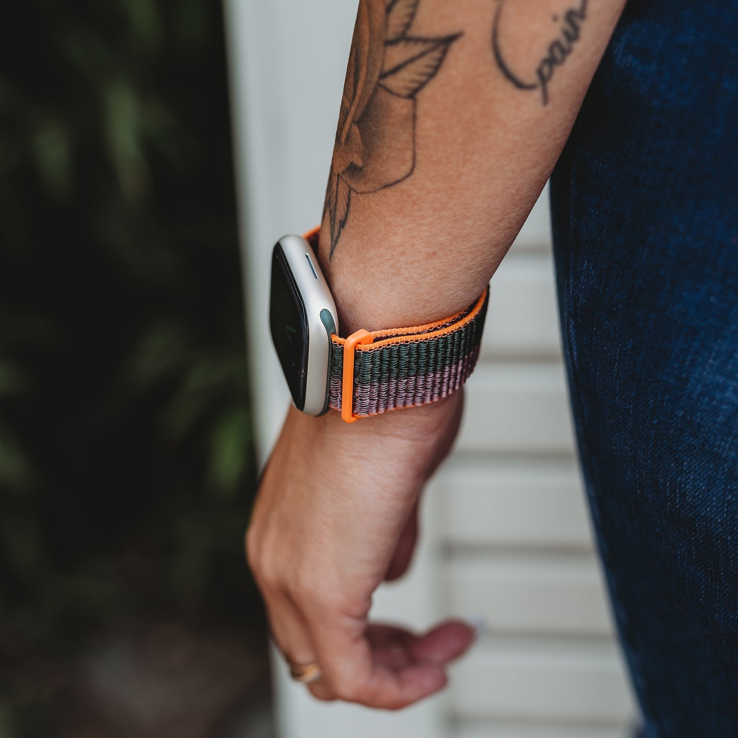 Draußen steht jemand mit dem arktisband Apple Watch Sportarmband „Fun“ mit seinem bunten gewebten Nylonband. Über seiner Bluejeans prangt vor einer weißen Wand ein Blumentattoo.