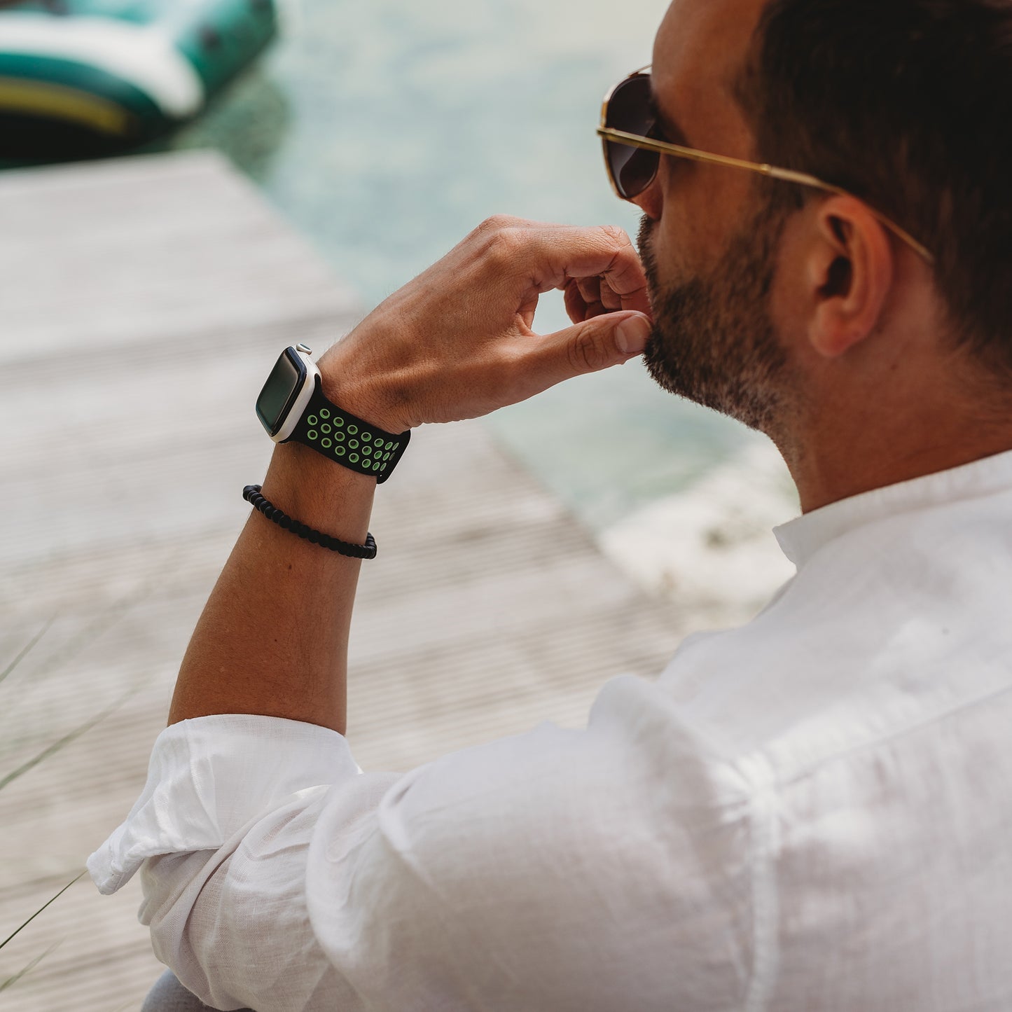 Ein Mann in weißem Hemd und Sonnenbrille sitzt auf einem Holzsteg am Wasser und trägt ein arktisband Apple Watch Silikonarmband „Energy“ von arktisband mit einem grünen perforierten Band und einem schwarzen Perlenarmband.