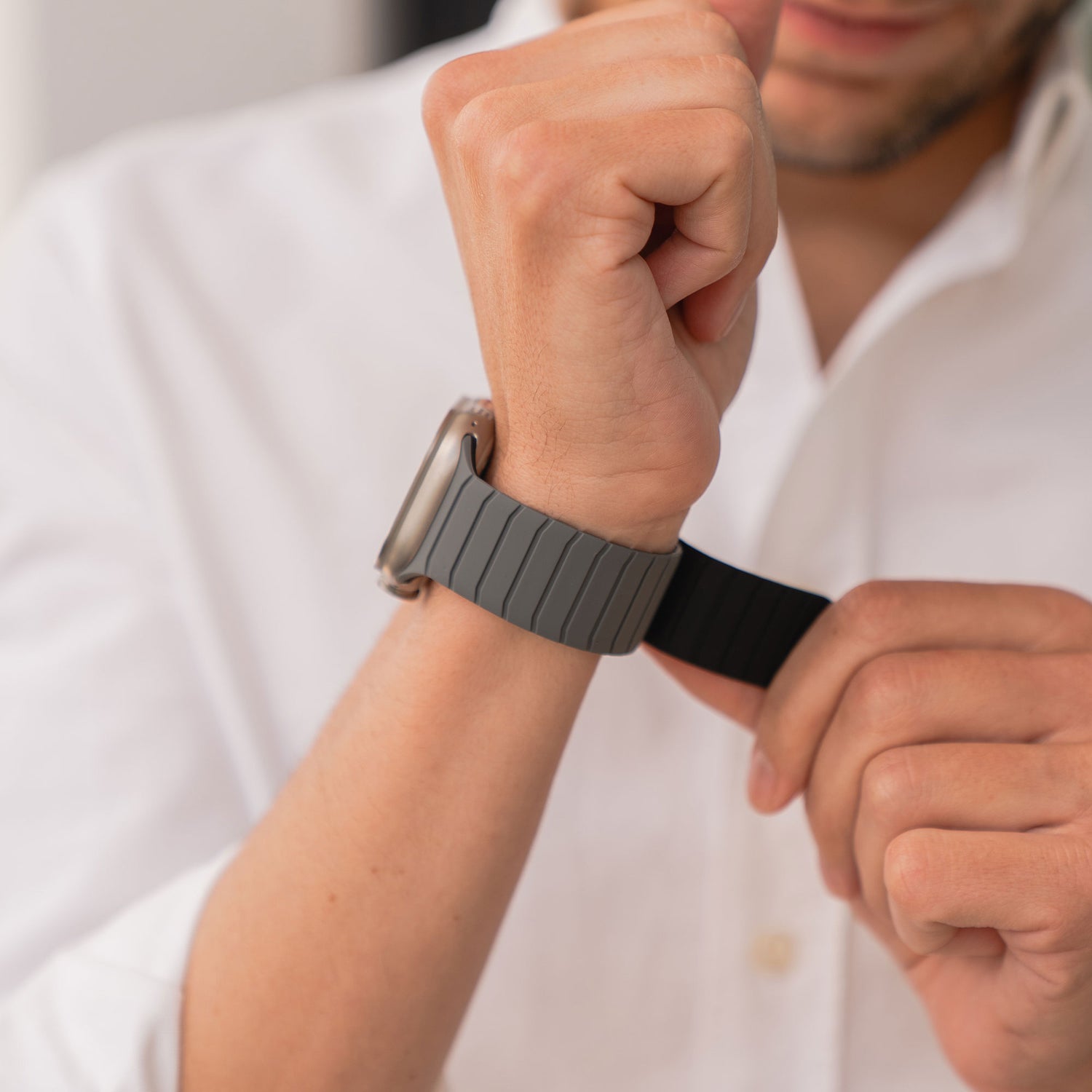 Eine Person im weißen Shirt legt das magnetische Armband „MagFlow“ aus Silikon mit Magnetverschluss an ihr linkes Handgelenk und konzentriert sich darauf, die Uhr anzupassen. Ihr Gesicht ist teilweise sichtbar.