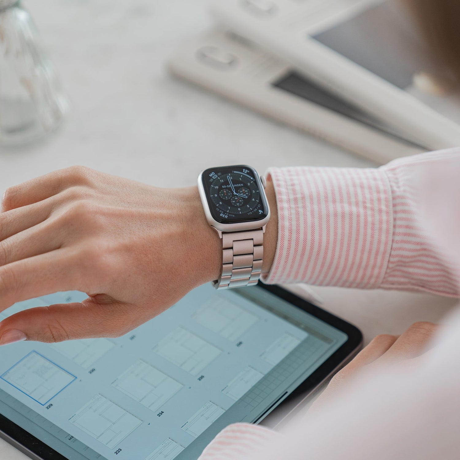 Eine Person, die das arktisband Apple Watch Gliederarmband „Arctica“ von arktisband trägt, berührt ein Tablet, auf dem Dokumente angezeigt werden, im Hintergrund sind auf einer weißen Fläche weitere elektronische Geräte zu sehen.