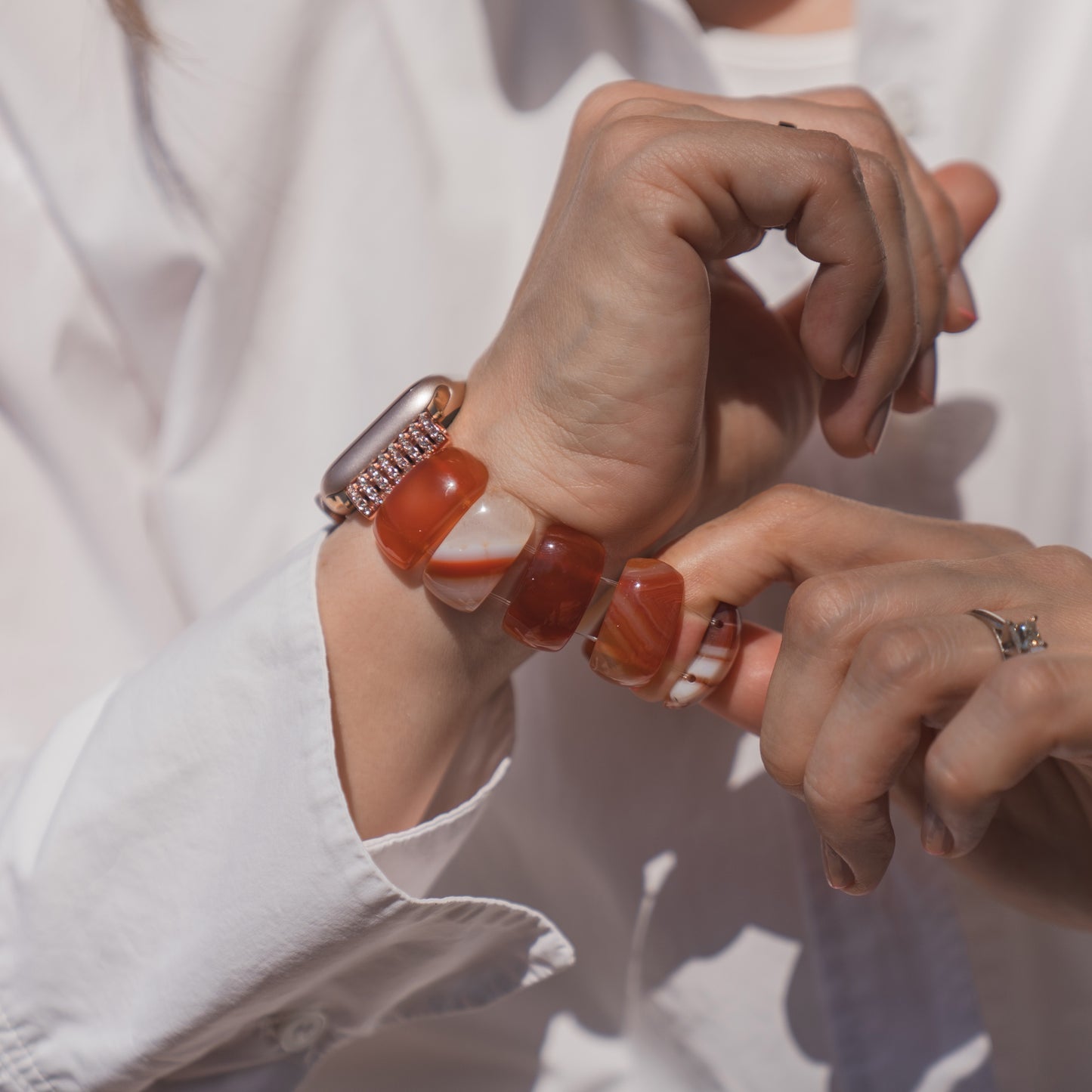 Eine Person in einem weißen Hemd berührt ihr Handgelenk; sie trägt ein rotes Arktisband für die Apple Watch „Stone Pearl“ und ein dazu passendes Naturstein-Armband.