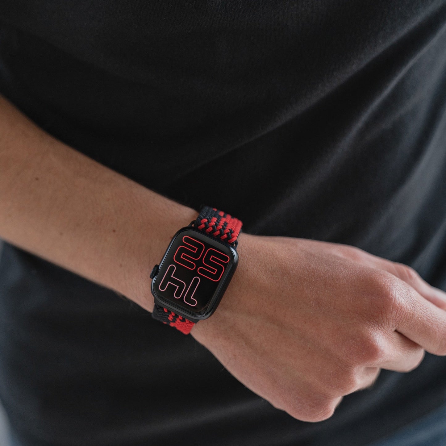 Eine Person in einem schwarzen Hemd trägt ein geflochtenes Flex Loop TwoTone Armband mit Edelstahlverschluss für die Apple Watch. Die Uhr zeigt große rote Ziffern: „25“ und „14“.