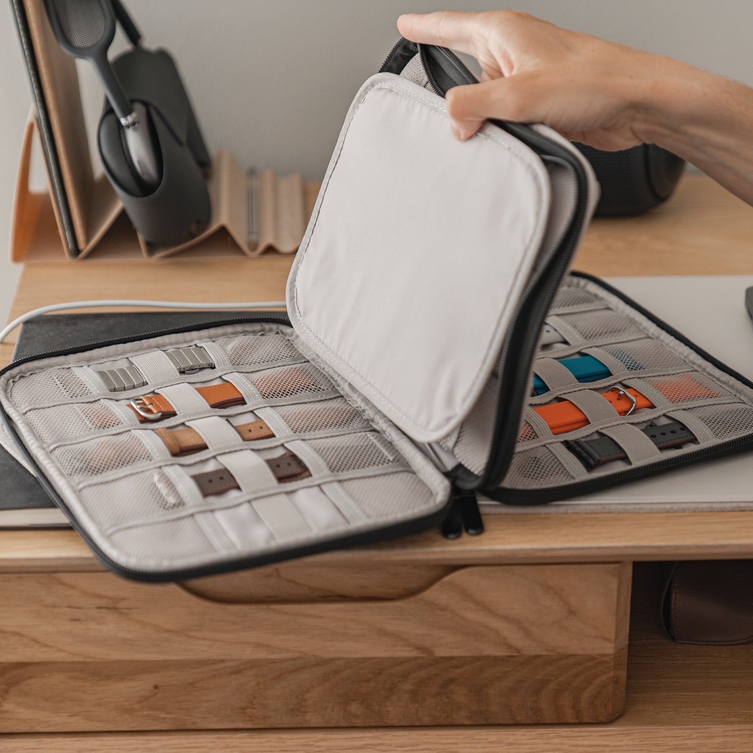 Eine Hand hält eine kleine Reisetasche für Apple Watch Armbänder & Zubehör von arktisband auf und zeigt ordentlich organisierte Armbänder in Netztaschen. Der Arktisband-Koffer steht auf einem Holzschreibtisch, daneben Büromaterial und Kopfhörer.