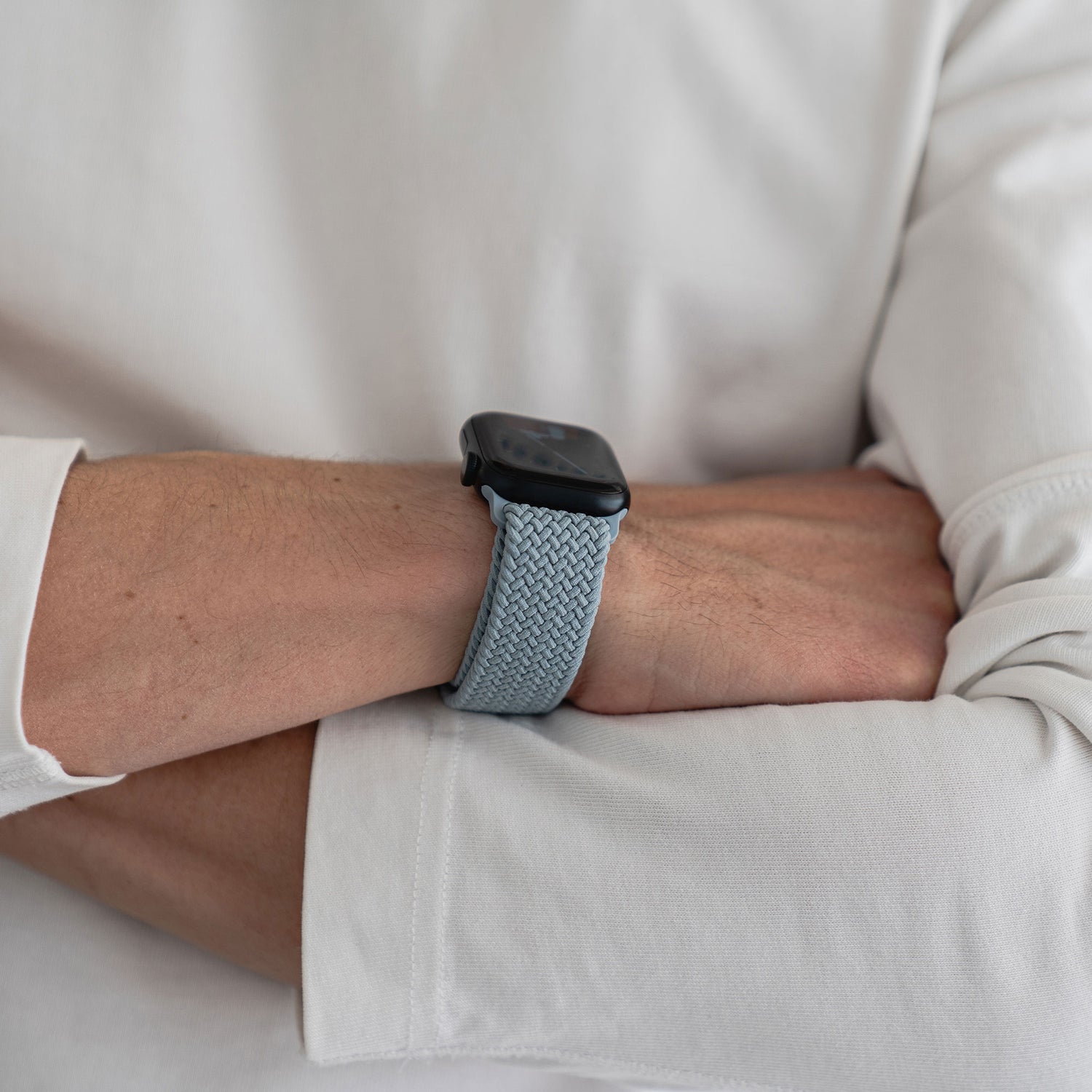 Person trägt eine dunkle Apple Watch mit einem Arktisband (Geflochtenes Apple Watch Armband „Casual“ mit Magnetverschluss) in Hellblau, die Arme sind verschränkt, sie trägt ein weißes Langarmshirt.