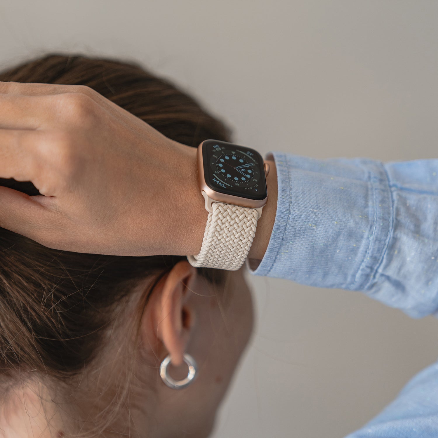 Eine braunhaarige Person mit Pferdeschwanz richtet den Kragen ihres blauen Hemdes und trägt ein geflochtenes Apple Watch Armband „Casual“ mit Magnetverschluss und silberne Creolen mit einem analogen Zifferblatt.