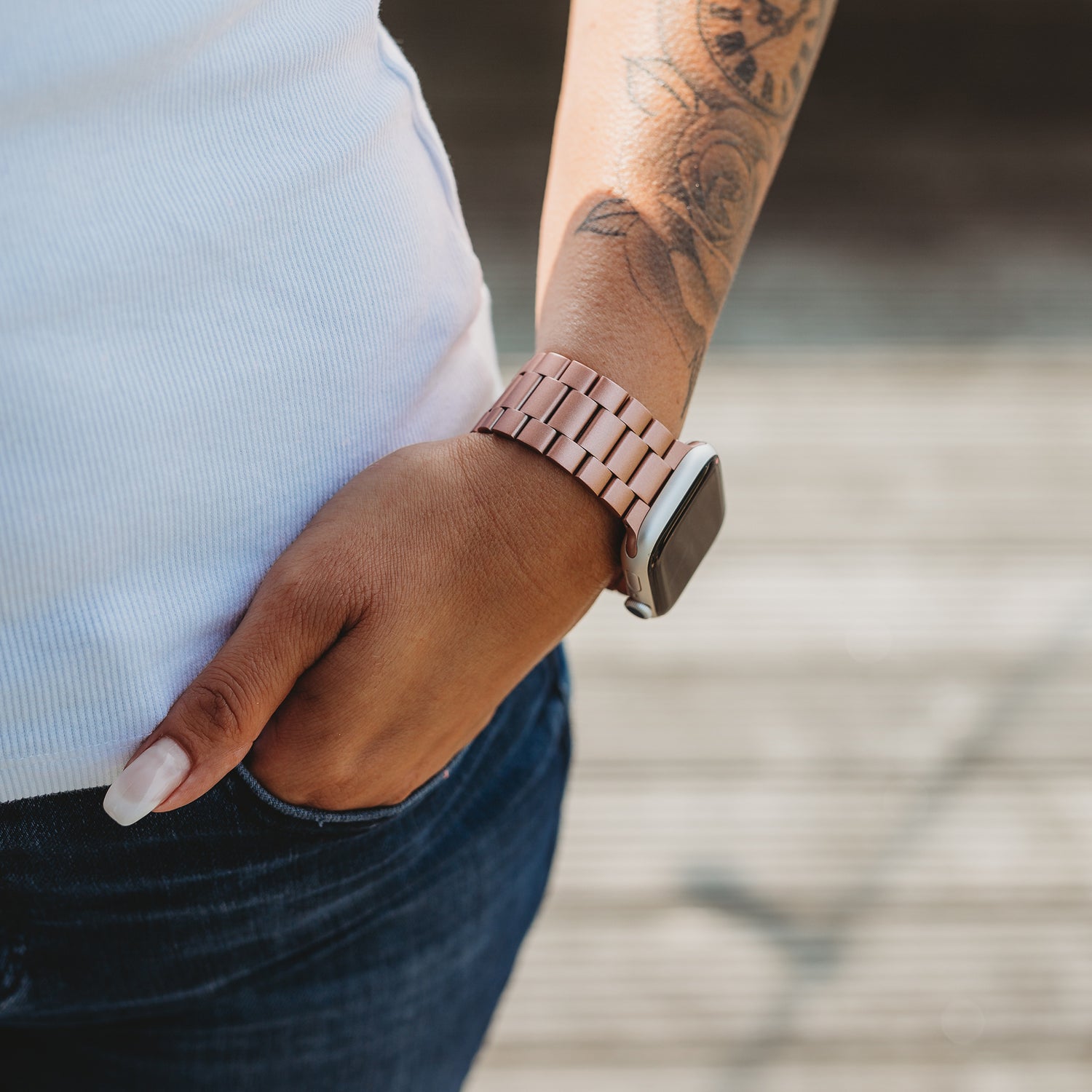 Eine Person, die ein Arktisband Apple Watch Gliederarmband „Arctica“ am linken Handgelenk trägt, steht mit einer Hand in der Jeanstasche da und gibt den Blick auf einen Teil eines weißen Hemdes und ein Blumentattoo auf dem Unterarm frei.