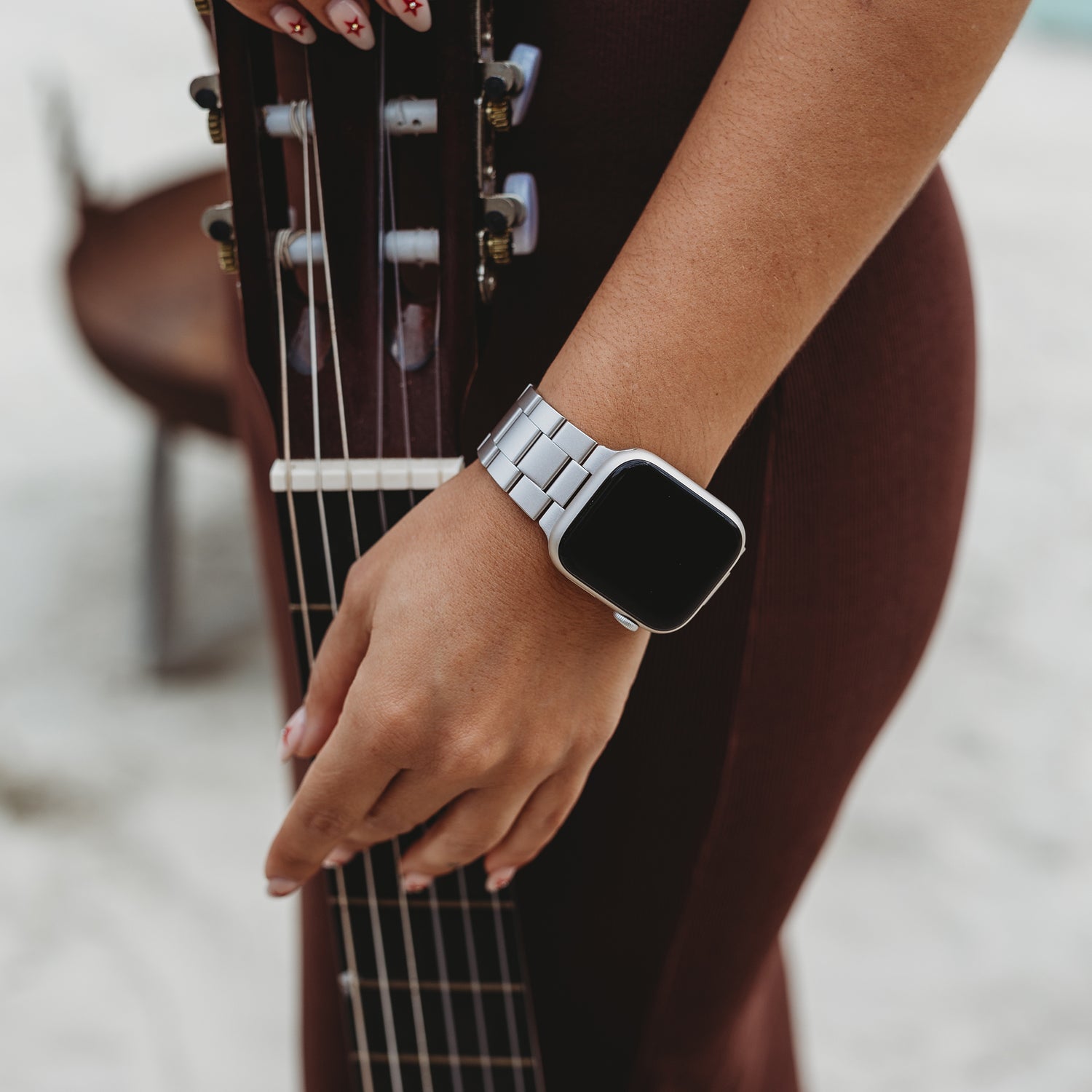 Eine Person in braunem Outfit mit gemusterten Nägeln trägt ein Arktisband Apple Watch Gliederarmband „Prime“ und hält eine Akustikgitarre im Freien, möglicherweise in einem sandigen Gebiet.