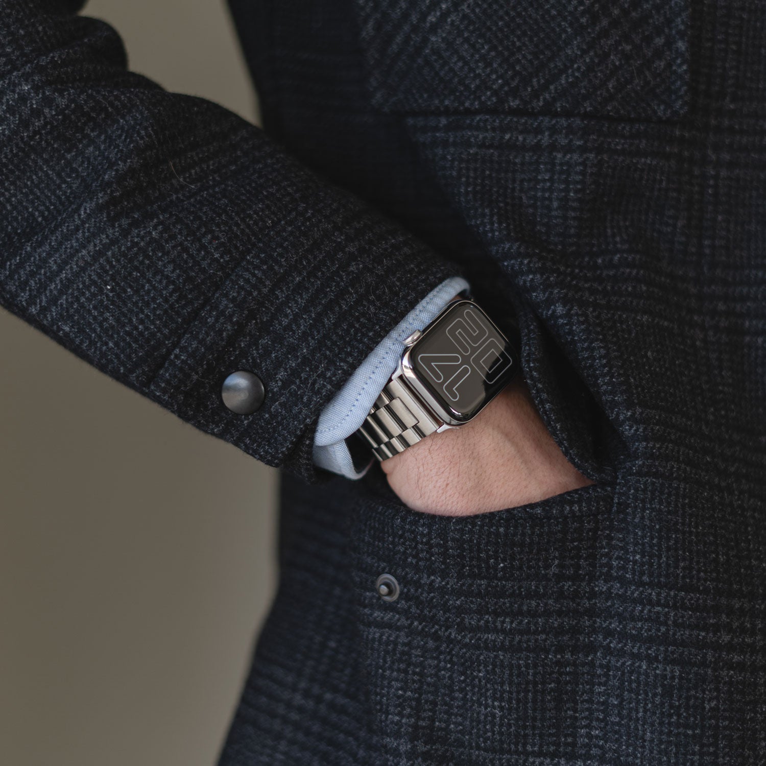 Eine Person in einem dunkelkarierten Mantel mit der linken Hand in der Tasche trägt ein arktisband Apple Watch Gliederarmband „Arctica“ von arktisband, das über ein schlankes, rechteckiges Zifferblatt und ein elegantes Edelstahlarmband verfügt.