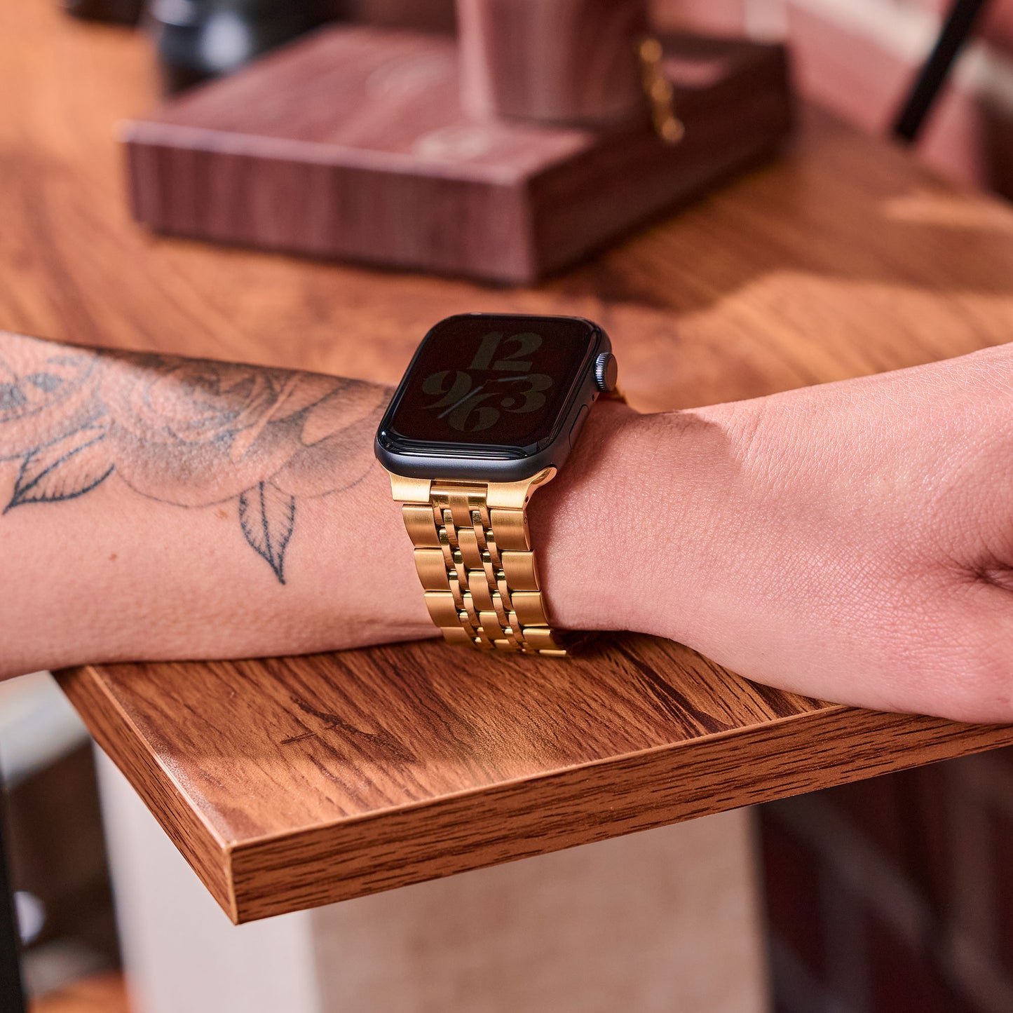 Eine Person mit floralem Unterarmtattoo trägt das arktisband Apple Watch Gliederarmband „Classic“ mit goldenem Edelstahlband und Butterfly-Verschluss. Ihr Arm ruht auf Holz, auf dem Display der Uhr sind große, stilisierte Ziffern zu sehen.