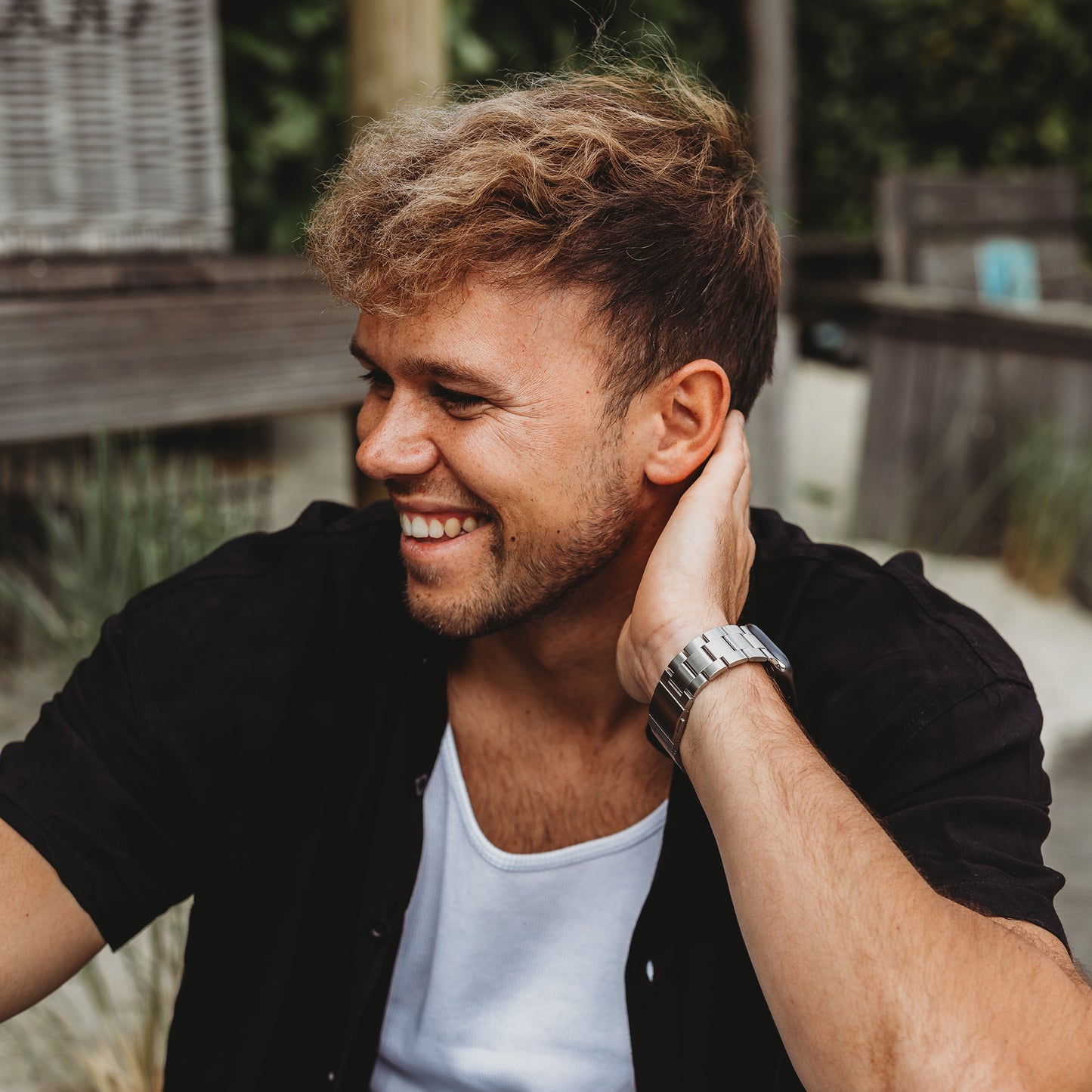 Ein lächelnder Mann mit hellbraunem Haar und kurzem Bart, der ein schwarzes Hemd, ein weißes T-Shirt und das Arktisband Apple Watch Gliederarmband „Prime“ trägt, berührt seinen Hals in einer hölzernen Außenkulisse.