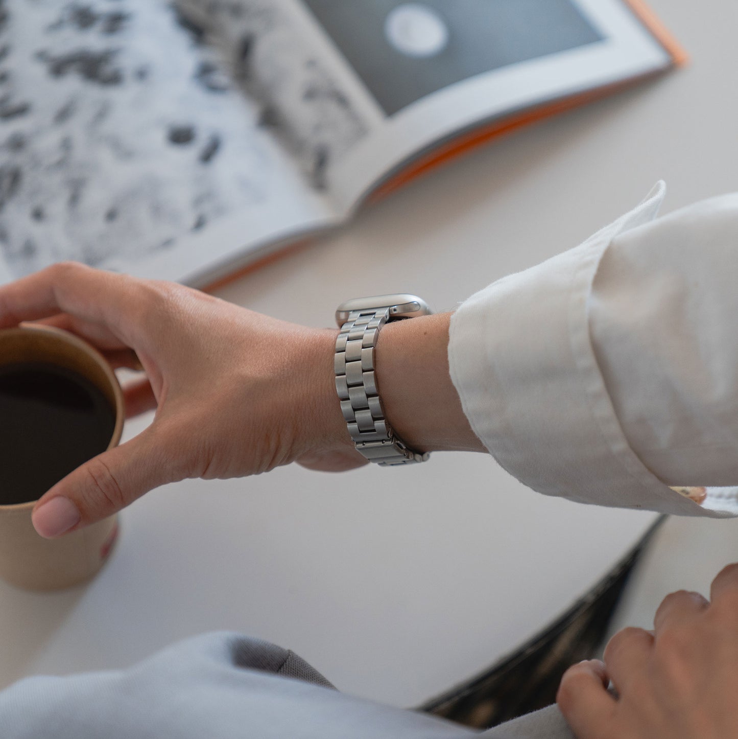 Eine Person mit dem Arktisband Apple Watch Gliederarmband "Arctica" - Slim und weißem Hemdsärmel hält einen Pappkaffeebecher. Im Hintergrund liegt auf einer weißen Fläche ein aufgeschlagenes, unscharfes schwarz-weißes Buch.