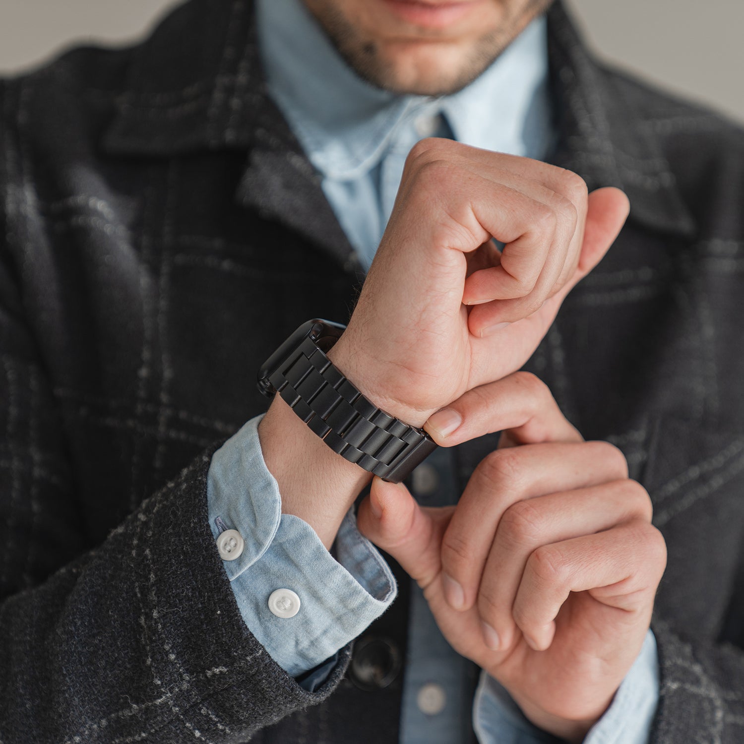 Eine Person in einer dunkelkarierten Jacke und einem hellblauen Hemd richtet ein Arktisband Apple Watch Gliederarmband „Arctica“ an ihrem linken Handgelenk, ihr Gesicht ist teilweise sichtbar und auf ihre Hände gerichtet.