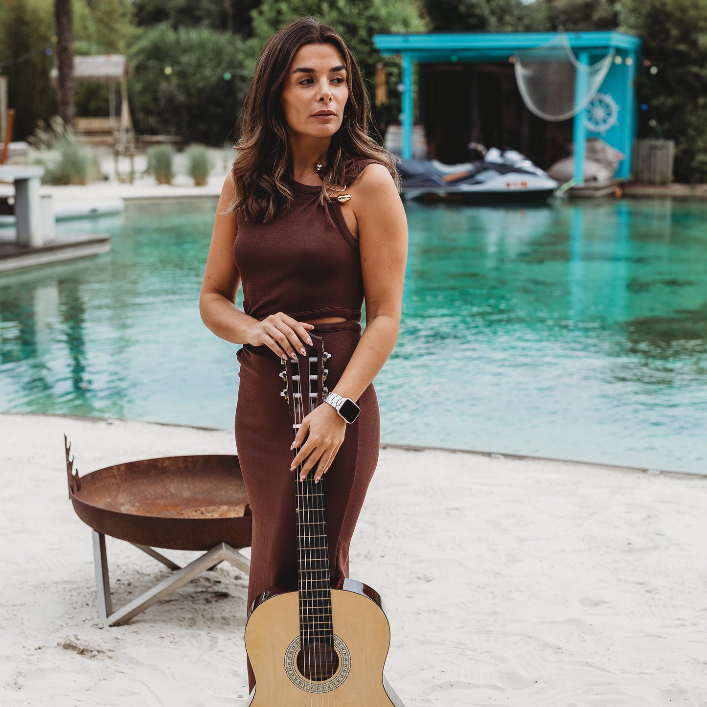 Eine Frau in einem ärmellosen braunen Kleid steht im Sand an einem Pool und hält eine Akustikgitarre aufrecht. Sie trägt das arktisband Apple Watch Gliederarmband „Prime“ von arktisband in der Nähe einer Feuerstelle. Hinter ihr sind eine blaue Struktur und ein Jetski zu sehen.