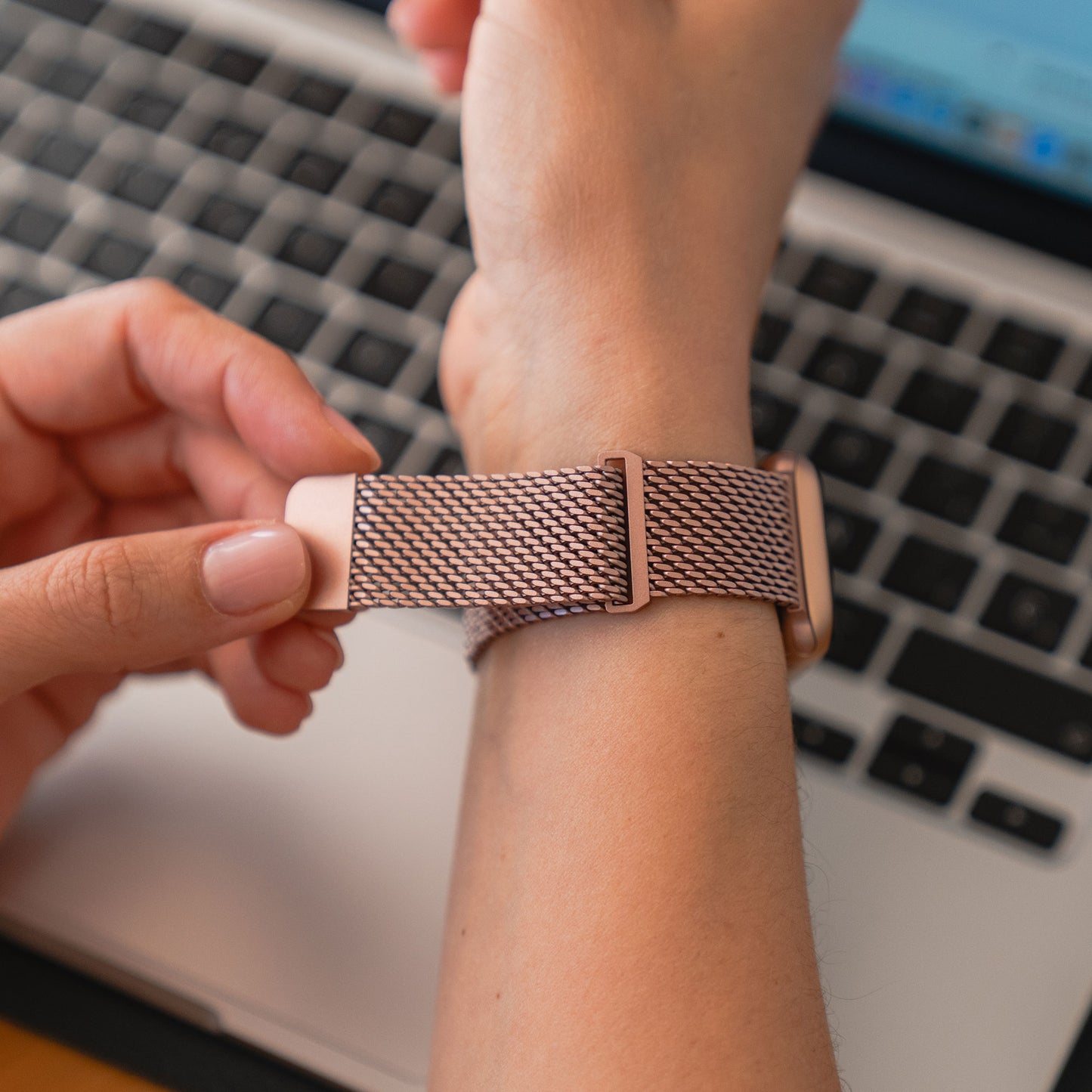 Eine Person befestigt das Arktisband Apple Watch Milanaise Armband „Bold“ in Roségold am Handgelenk, im Hintergrund ist eine Laptoptastatur zu sehen.