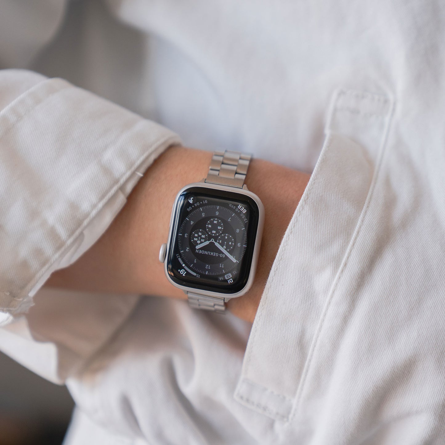 Eine Person trägt eine silberne Apple Watch mit dem Arktisband Apple Watch Gliederarmband „Arctica“ – Slim. Das schwarze Ziffernblatt im Analogstil ist teilweise unter einem weißen Jackenärmel sichtbar. Das elegante Arktisband-Accessoire verleiht der Uhr einen modernen, stilvollen Touch.