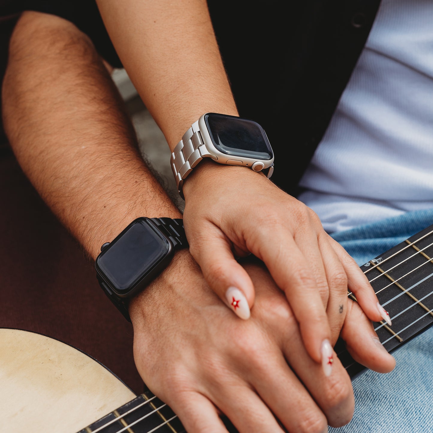 Zwei Personen mit Smartwatches – eine trägt ein arktisband Apple Watch Gliederarmband „Prime“ in Silber, die andere ein schwarzes Armband – stützen ihre Hände auf einer Gitarre. Die Fingernägel der oberen Person sind mit einem roten Sternmotiv verziert.