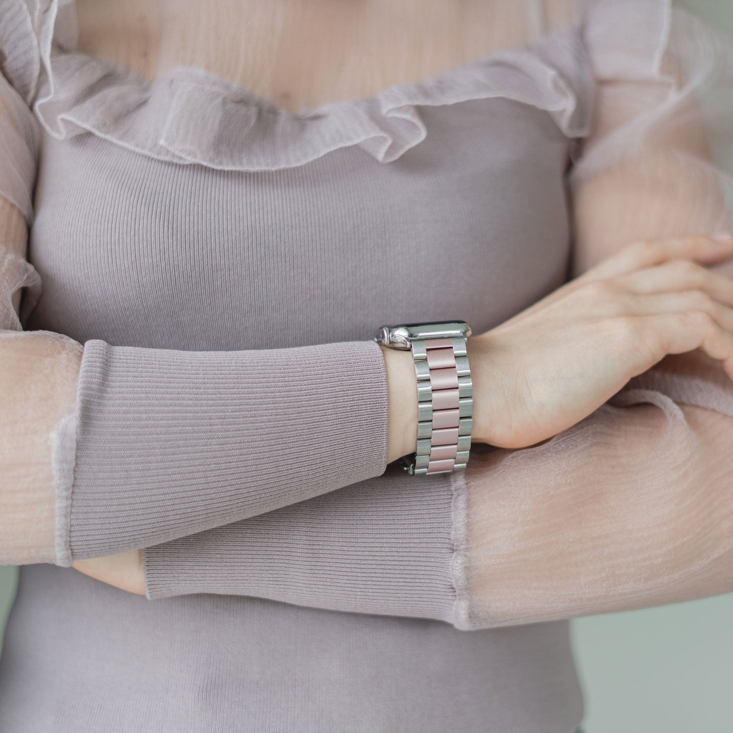 Eine Person in einem transparenten, langärmligen lavendelfarbenen Oberteil verschränkt die Arme und zeigt ein elegantes Arktisband Apple Watch Gliederarmband „Arctica“ mit abwechselnd silbernen und rosa Gliedern am Handgelenk.