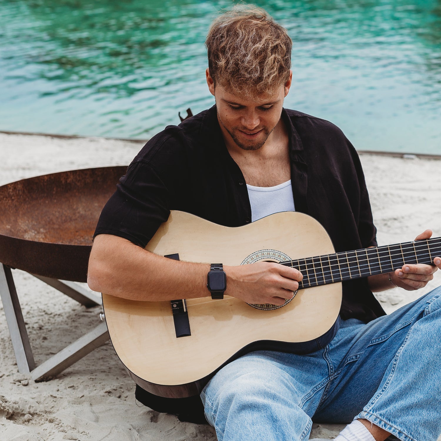 Ein Mann in einem schwarzen Hemd über einem weißen Top und Blue Jeans spielt Akustikgitarre an einer Feuerstelle und am Wasser. Er trägt das arktisband Apple Watch Gliederarmband „Prime“ von arktisband und blickt auf seine Gitarre hinunter.