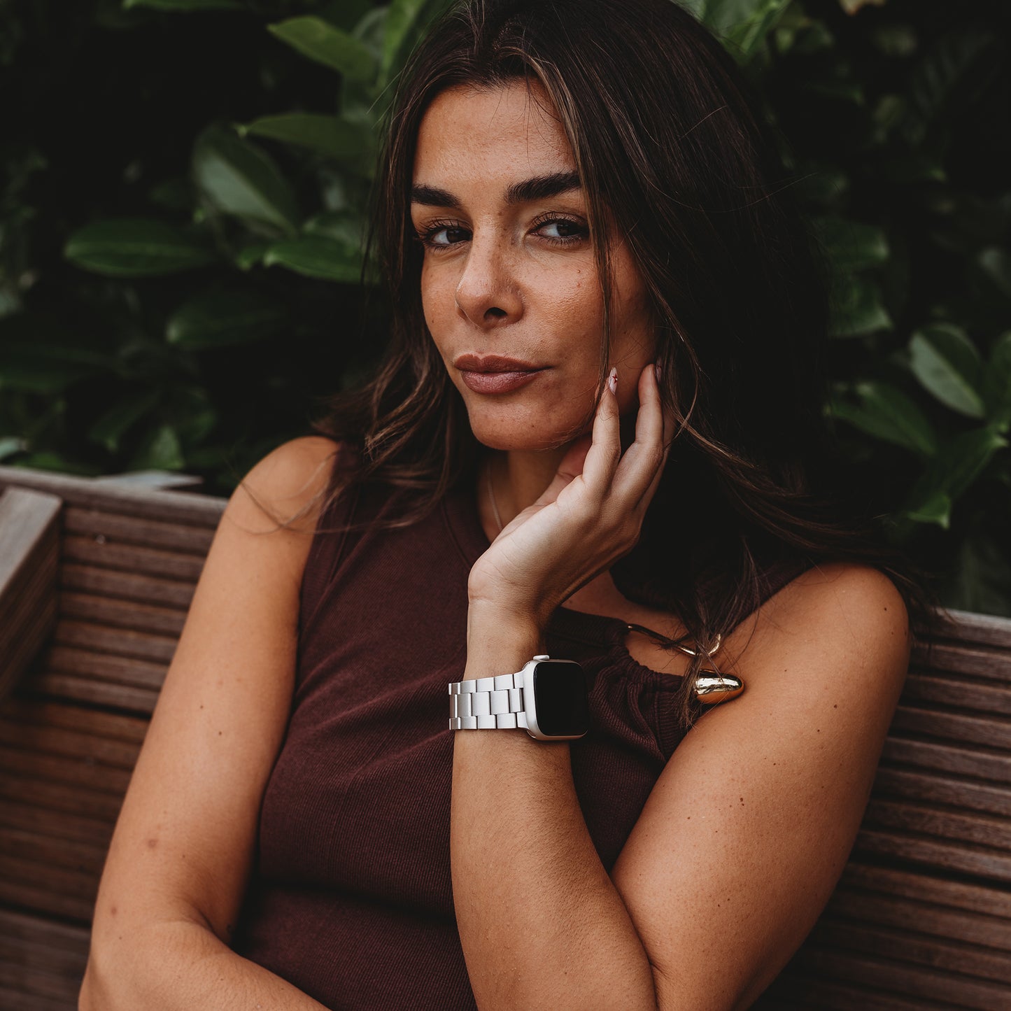 Eine Frau mit langen braunen Haaren sitzt auf einer Holzbank. Sie trägt ein ärmelloses braunes Top und das arktisband Apple Watch Gliederarmband „Prime“ von arktisband. Sie blickt in die Kamera, das Kinn auf der Hand, im Hintergrund grünes Laub.