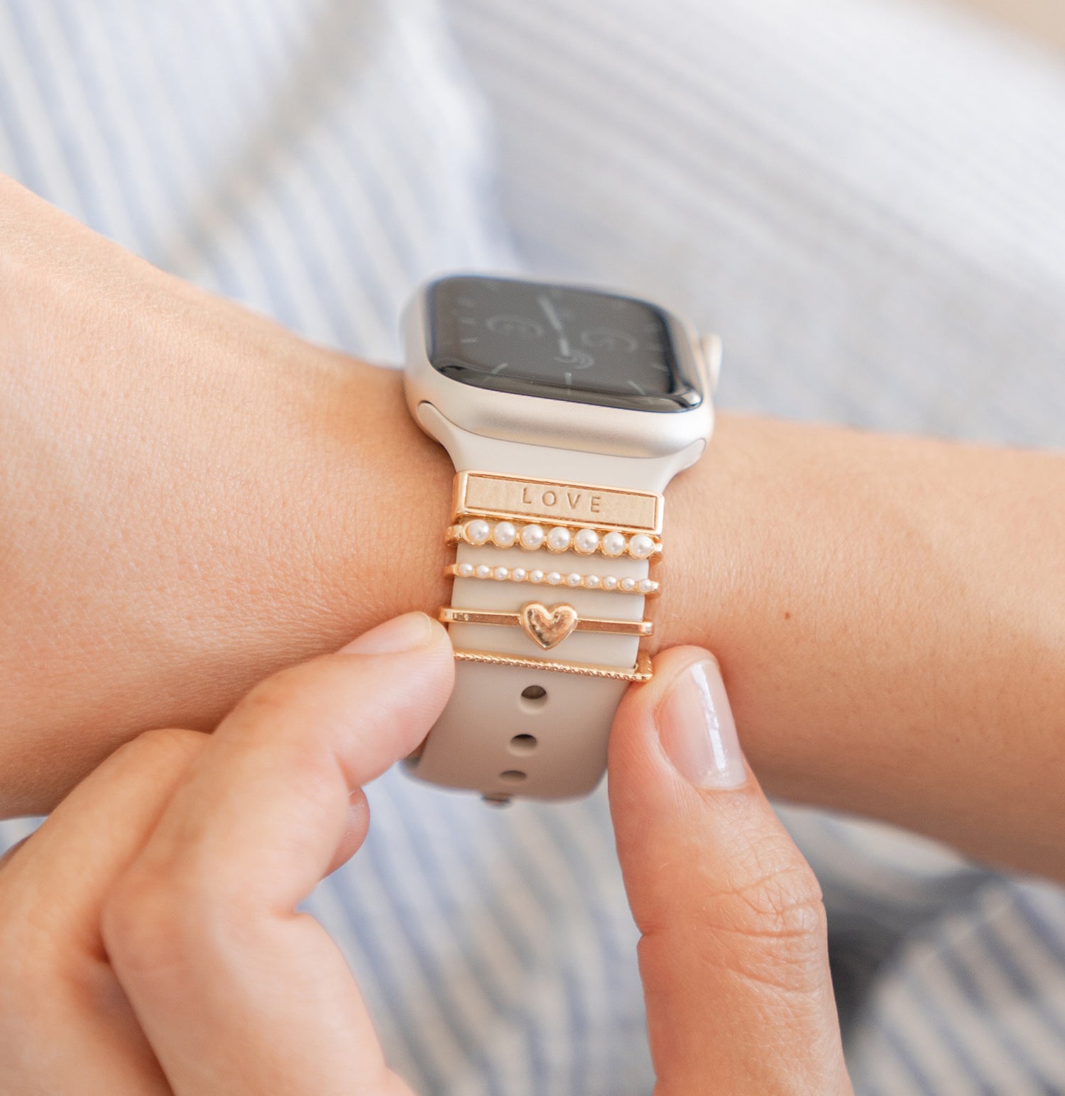 Eine Hand trägt eine Smartwatch mit Anhängern aus Gold und Perlen am Armband, darunter einer mit der Gravur „LOVE“.
