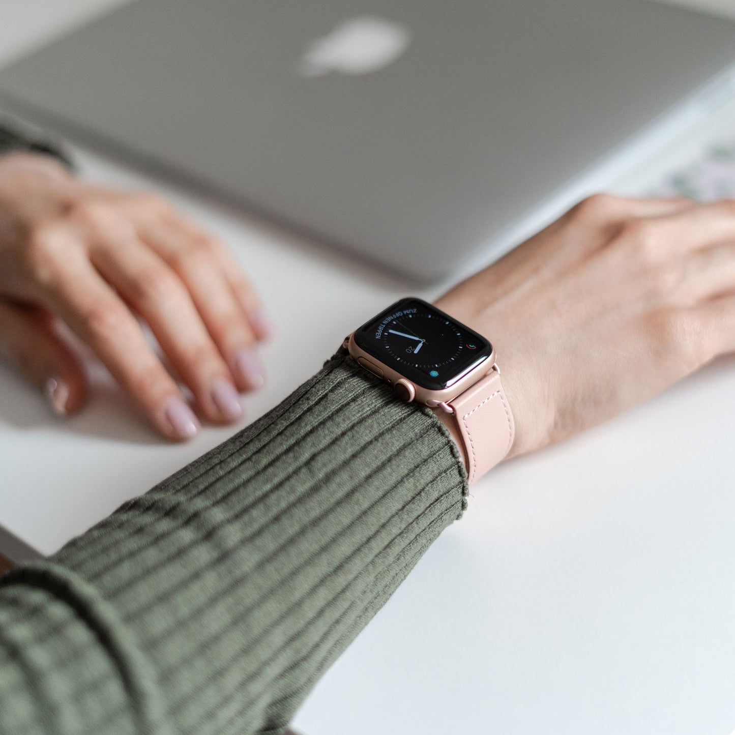 Eine Person trägt ein grünes, geripptes Langarmshirt und präsentiert einen minimalistischen Look mit dem Arktisband Apple Watch Lederarmband „Novus“ in Pink am linken Handgelenk neben einem geschlossenen silbernen Laptop auf einem weißen Schreibtisch.