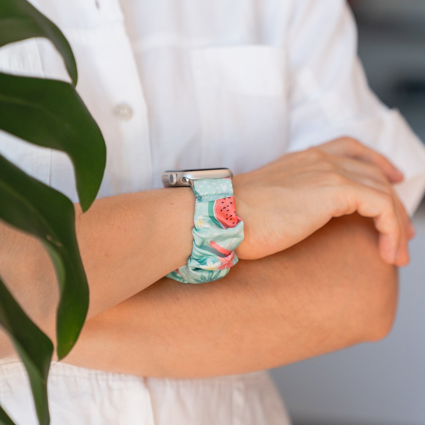 Eine Person trägt das arktisband Apple Watch Scrunchie Armband "Elanora" von arktisband mit einem farbenfrohen, geblümten Gummiband mit einem kleinen Wassermelonen-Anhänger und steht mit verschränkten Armen.