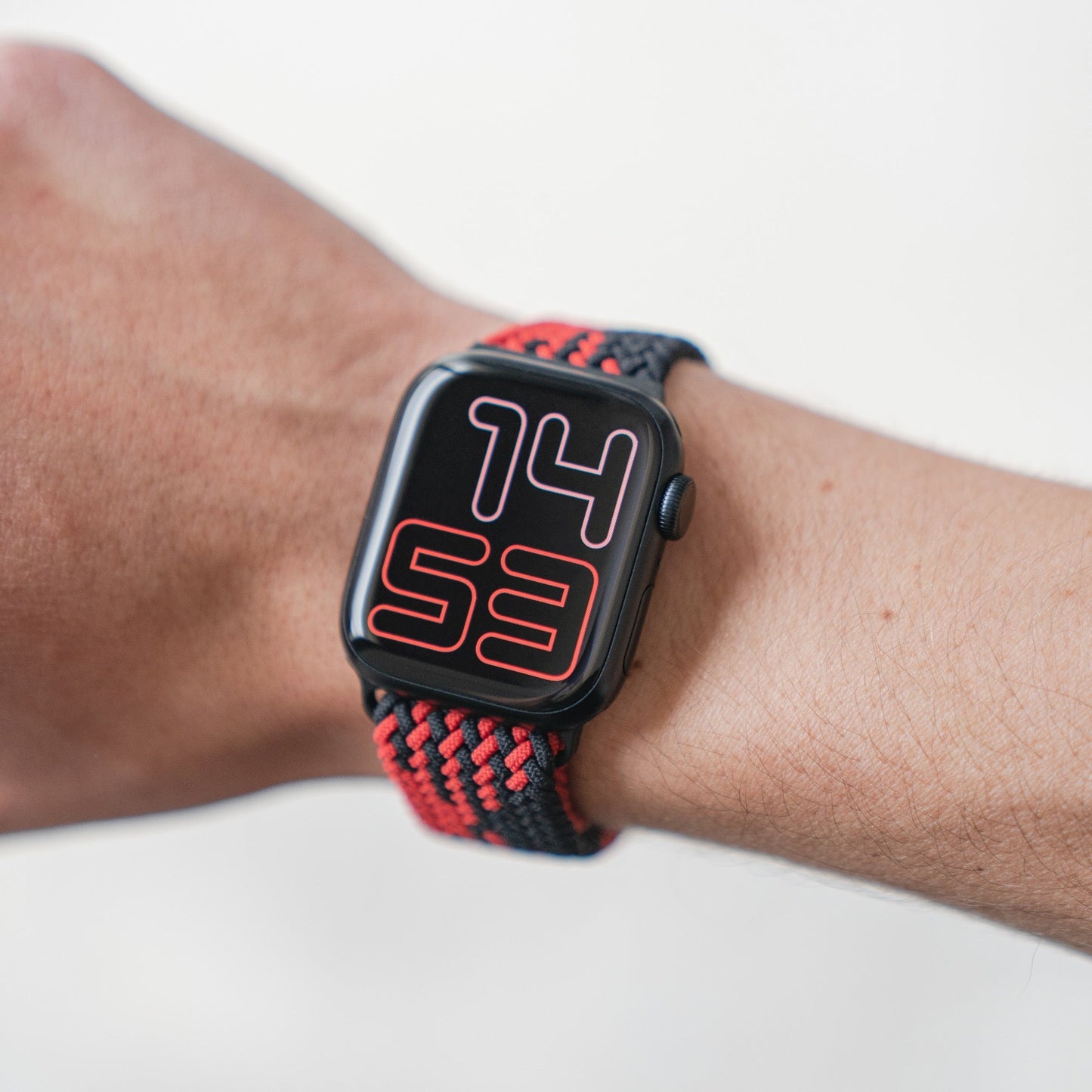 Am Handgelenk wird eine Arktisband Apple Watch mit schwarzem Gehäuse und geflochtenem Flex Loop TwoTone Armband in Schwarz/Rot getragen, das durch einen Edelstahlverschluss gesichert wird. Ihre Digitalanzeige zeigt 14:53 in großen rosa und roten Ziffern an.