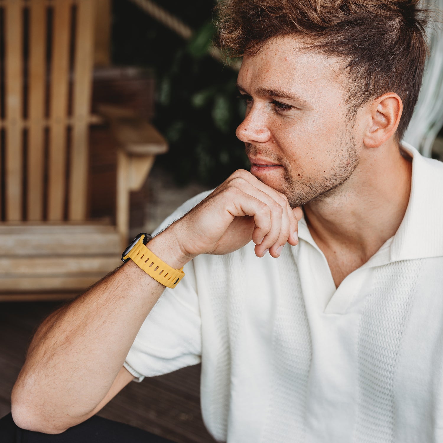 Ein Mann mit kurzen braunen Haaren und Bart sitzt im Freien, trägt ein weißes strukturiertes Poloshirt und eine Apple Watch Ultra mit dem Arktisband „Active“ Silikonarmband in Gelb, stützt sein Kinn auf seine Hand und blickt zur Seite.