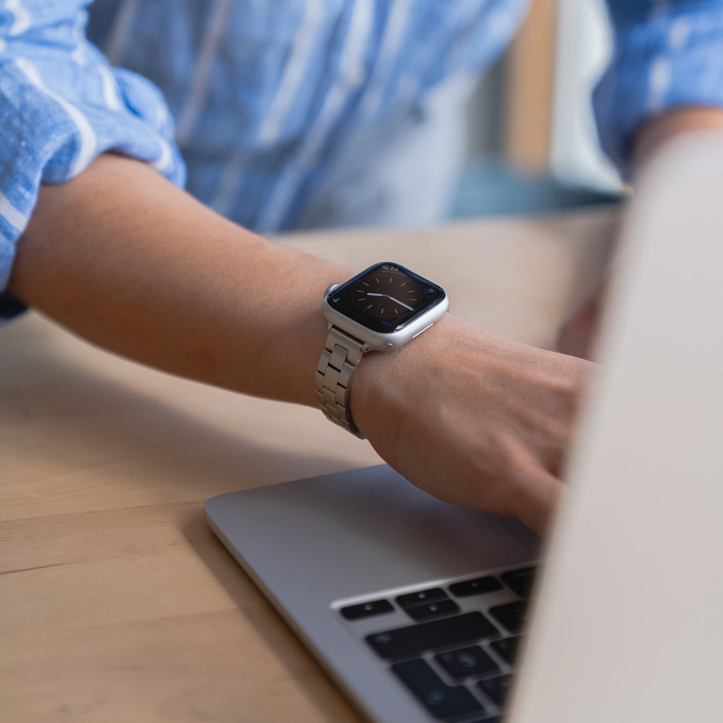 Eine Person im blauen Hemd arbeitet an einem Laptop an einem Holzschreibtisch und trägt das arktisband Apple Watch Gliederarmband „Arctica“ – Slim von arktisband.