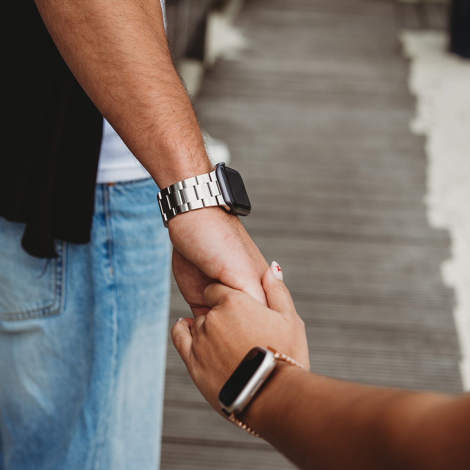 Zwei Personen halten sich auf einem Holzweg an den Händen. Beide tragen ein Arktisband Apple Watch Gliederarmband „Prime“. Eine trägt ein elegantes Edelstahlarmband mit MagneticClick-Verschluss, die andere ein klassisches schwarzes Armband. Nur ihre Arme und Hände sind sichtbar.