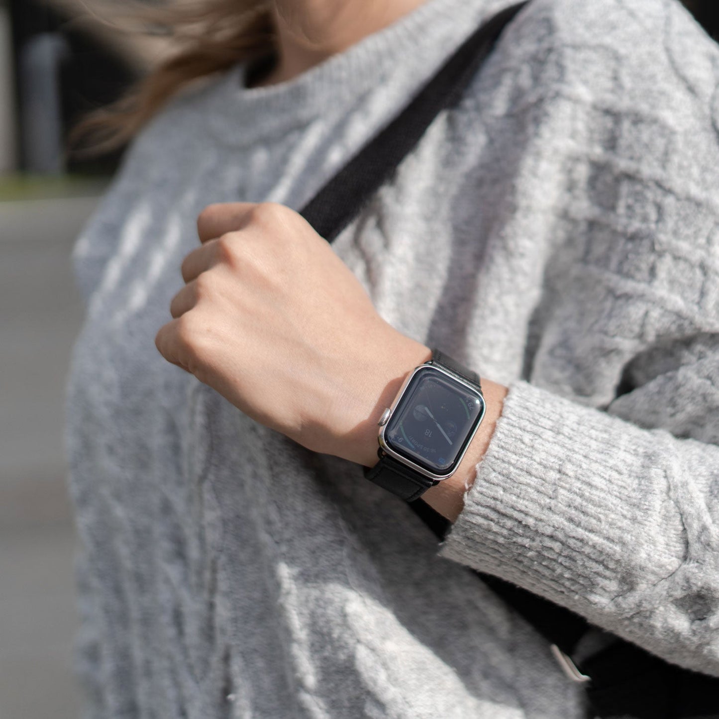 Person, die einen grauen Pullover und ein Arktisband Apple Watch Kunstlederarmband trägt und im Freien ein schwarzes Armband über der Schulter hält, um einen minimalistischen Look zu erzielen.