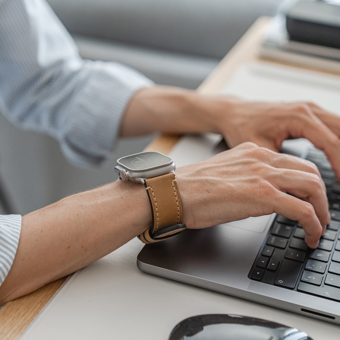Jemand trägt ein Arktisband Apple Watch Ultra PALERMO GRANDE Lederarmband und tippt an einem Schreibtisch auf einem Laptop. Eine Maus und ein Stapel Notizbücher oder Papiere liegen in der Nähe. Die Person trägt ein hellblau gestreiftes Hemd.