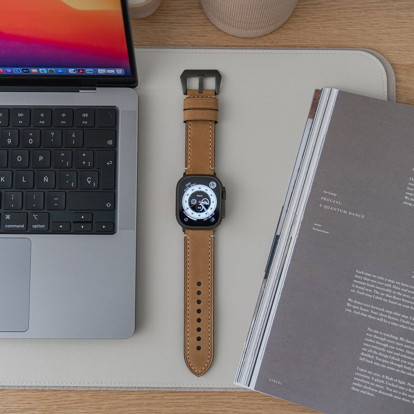 Eine arktisband Apple Watch Ultra mit einem PALERMO GRANDE Lederarmband liegt auf einer Schreibtischunterlage zwischen einem aufgeklappten Laptop und einer Zeitschrift. Das Display zeigt eine digitale Analoguhr.