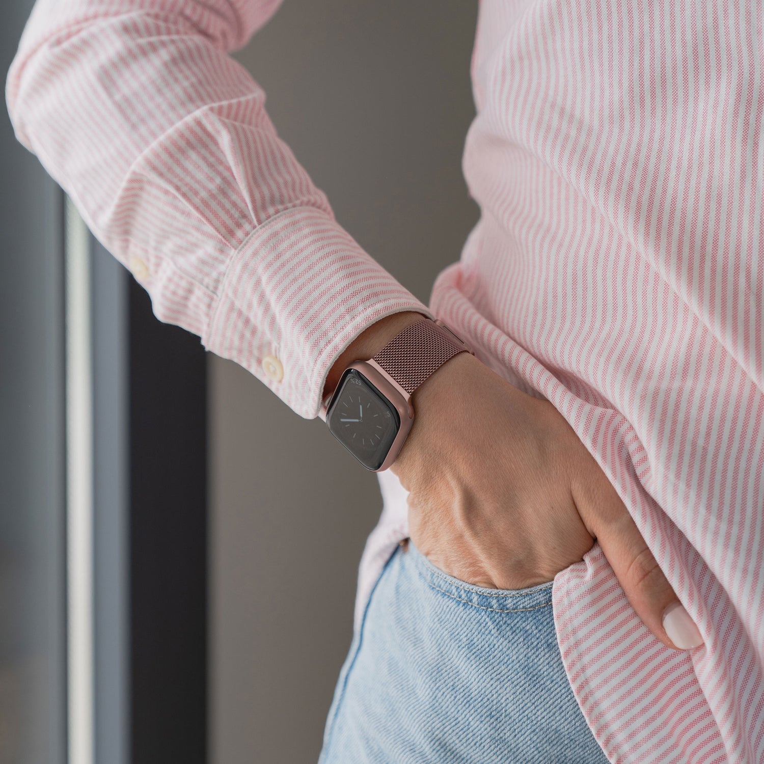 Eine Person in einem rosa-weiß gestreiften Hemd und hellblauen Jeans hat die Hand in der Tasche und zeigt ein arktisband Apple Watch Milanaise Loop Armband an ihrem Handgelenk.